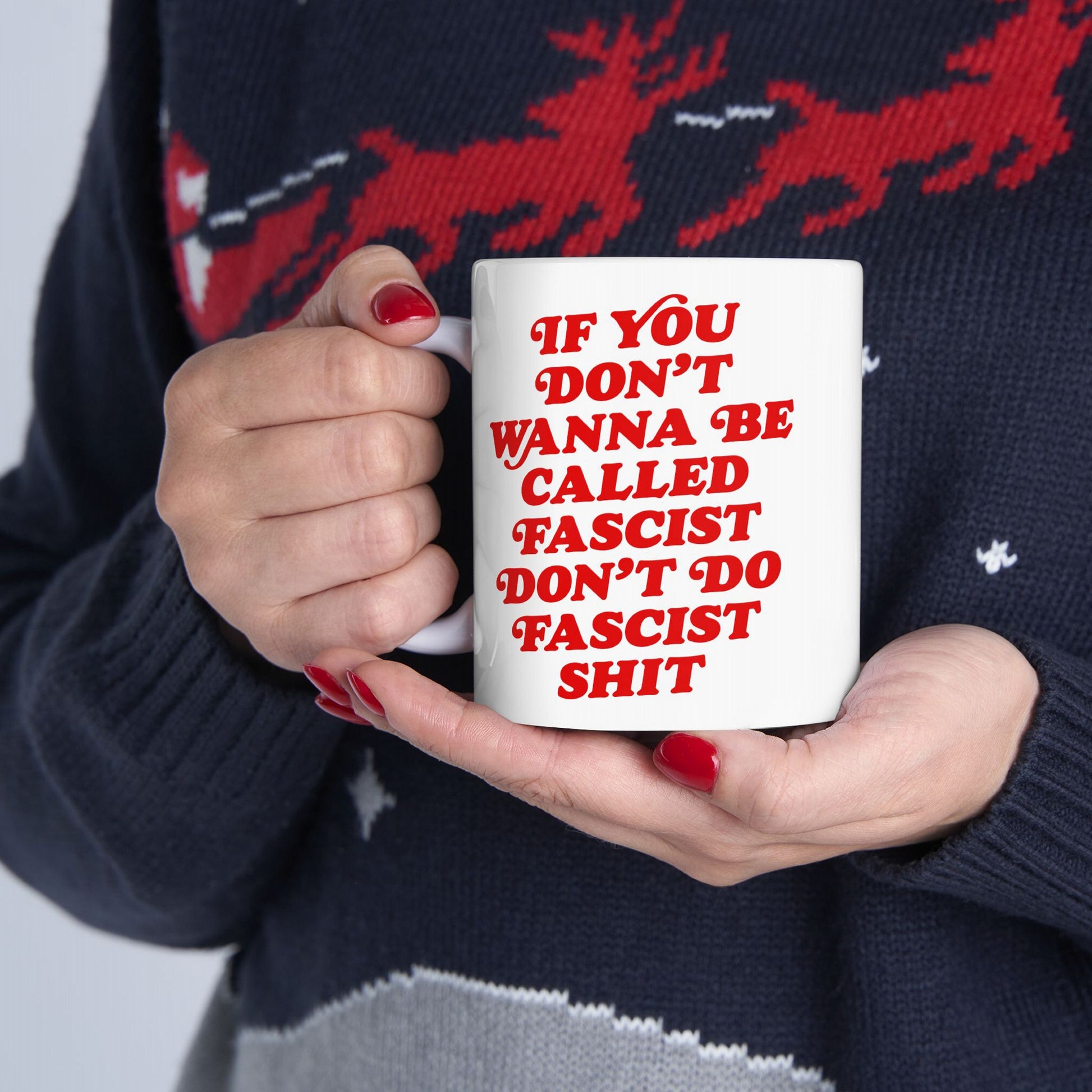 A person holds an Anti-Fascist Retro Mug with bold red lettering, featuring the slogan about resisting fascism. They wear a dark knit sweater, and their fingernails are painted red.