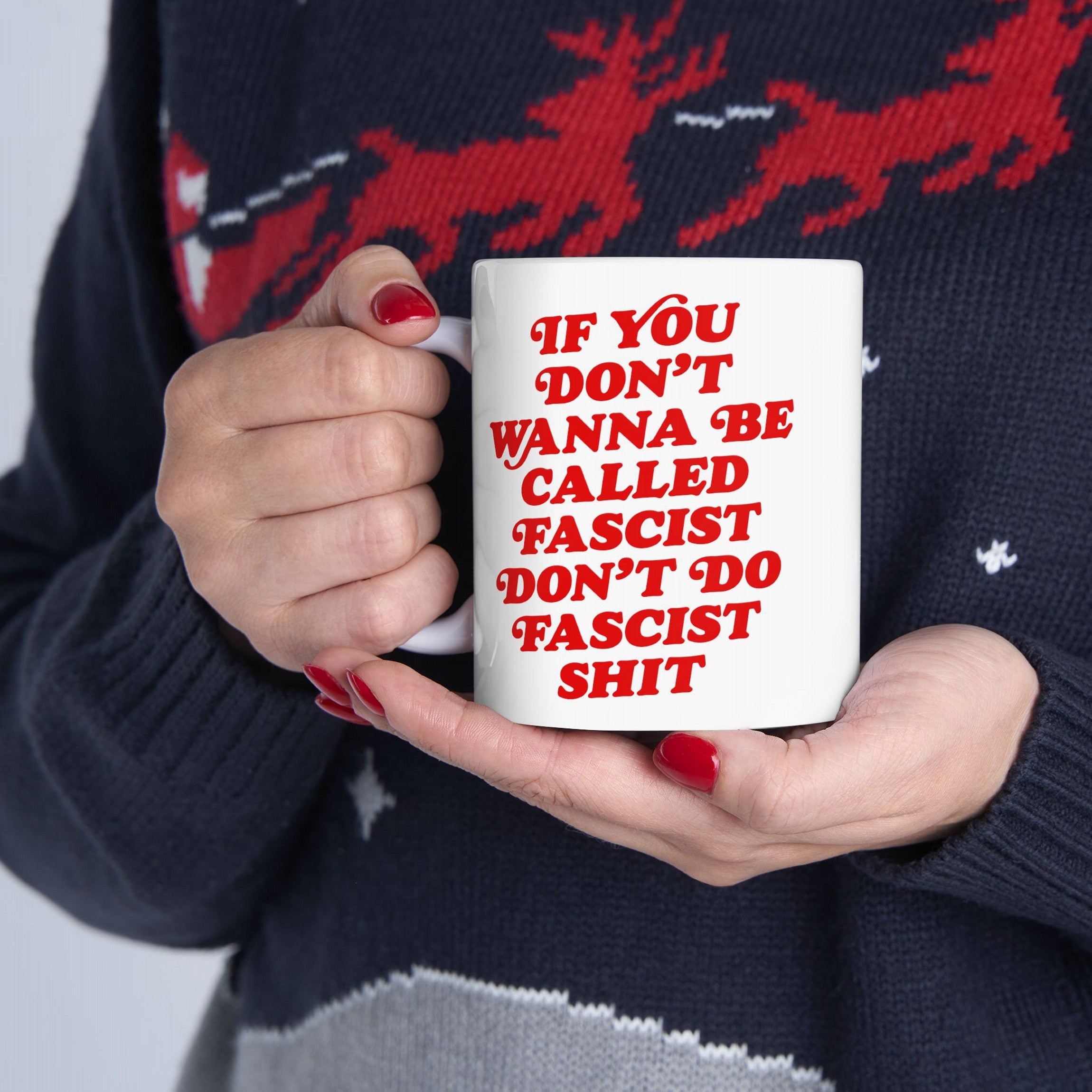A person holds an Anti-Fascist Retro Mug with bold red lettering, featuring the slogan about resisting fascism. They wear a dark knit sweater, and their fingernails are painted red.