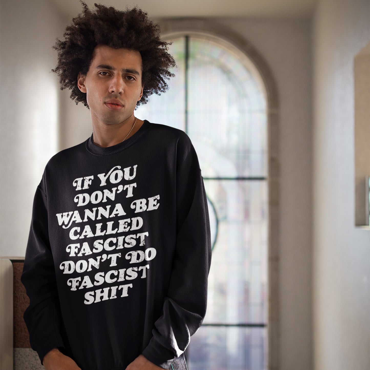 A young person wears an Anti-Fascist Retro Shirt, featuring the slogan If you don’t wanna be called fascist, don’t do fascist shit, standing before an arched window, embodying bold, silent resistance.