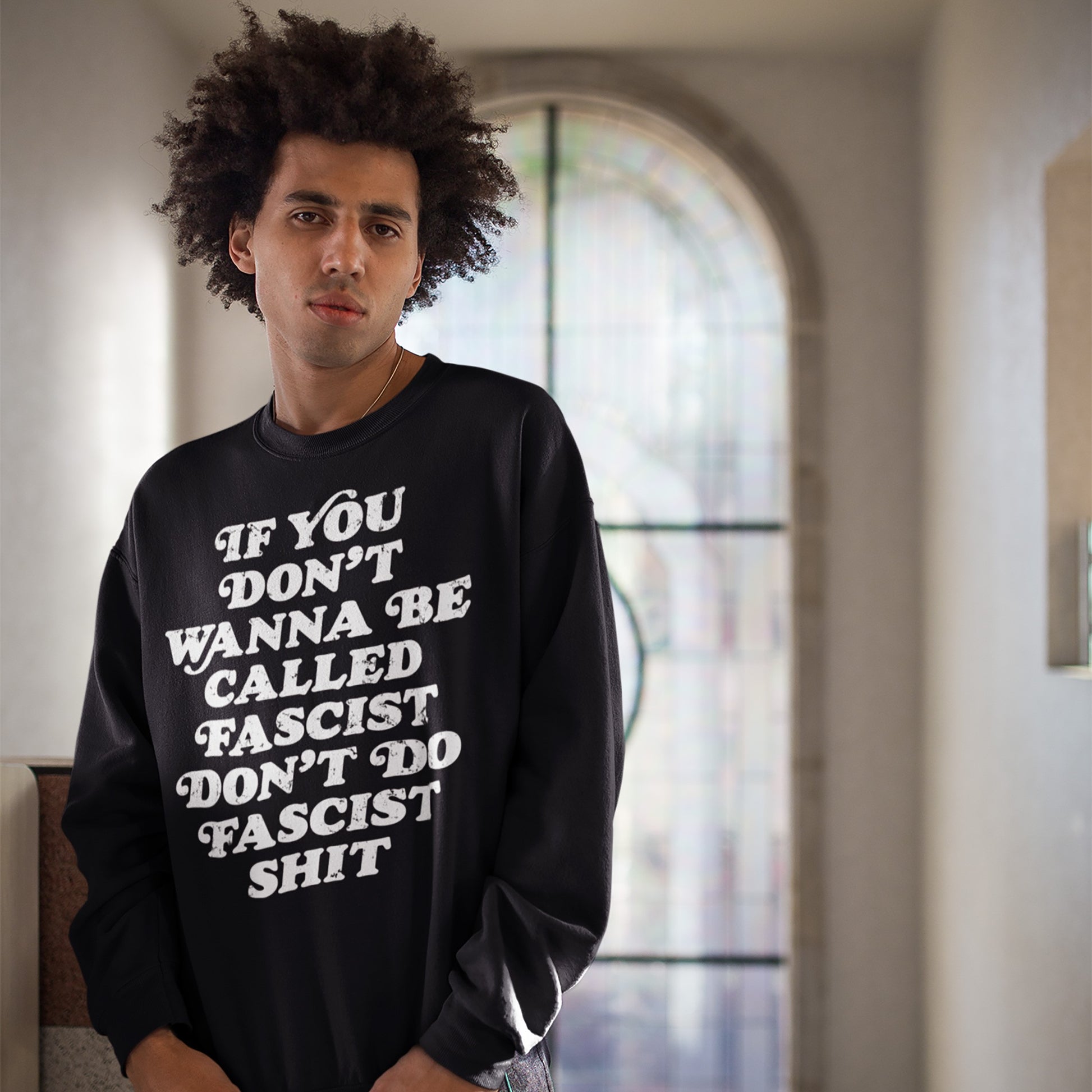 A young person wears an Anti-Fascist Retro Shirt, featuring the slogan If you don’t wanna be called fascist, don’t do fascist shit, standing before an arched window, embodying bold, silent resistance.