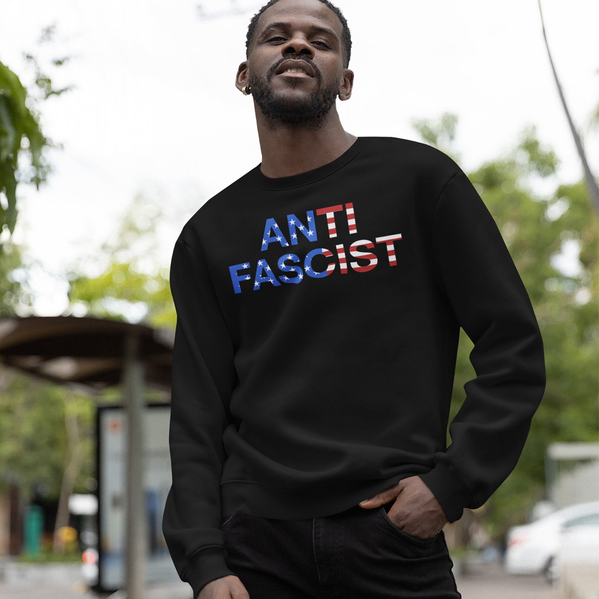 Man outdoors wearing the Anti-Fascist USA Classic Hoodie & Sweatshirt, featuring ANTI FASCIST text with American-flag motif, symbolizing a stand against tyranny and promoting activism and equality.
