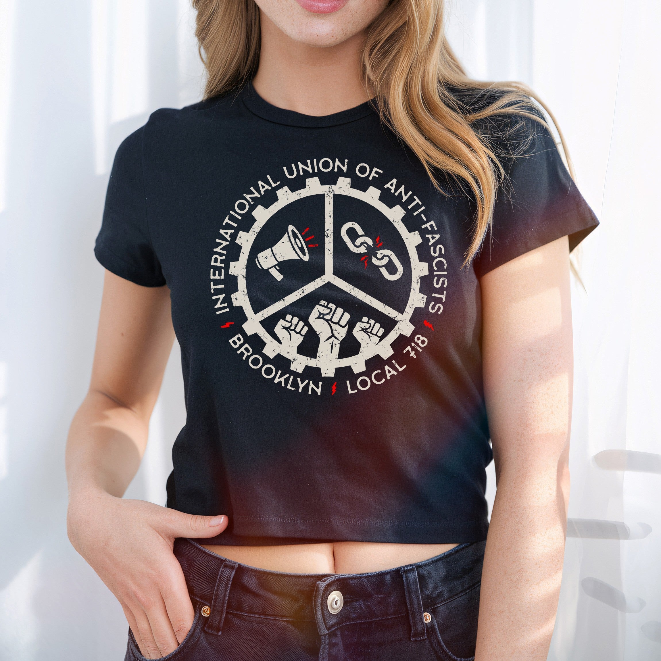Brooklyn Anti-Fascist Union Women's Baby Tee featuring a protest design with a circular gear logo showcasing a megaphone, broken chain, and raised fists, emphasizing anti-fascist solidarity and local identity.