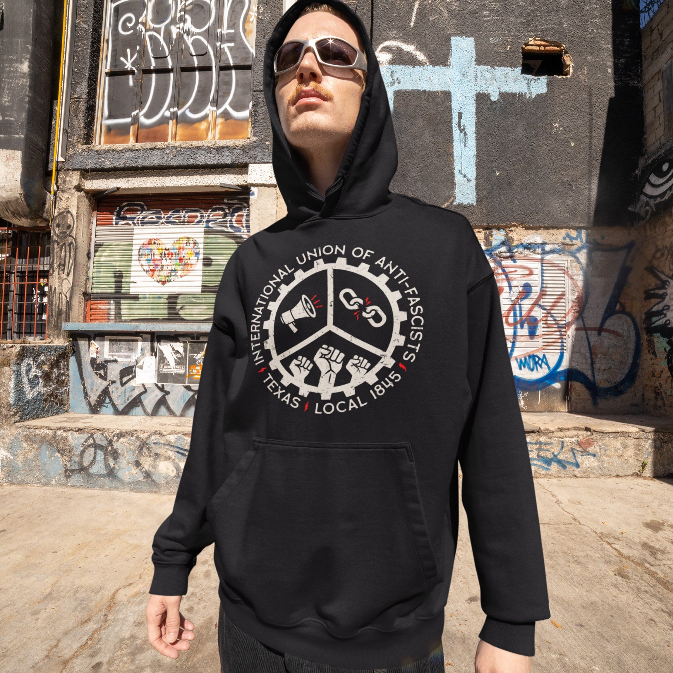 Person in urban setting wears Custom Anti-Fascist Union Hoodie featuring emblem with megaphone, broken chain, and fists, symbolizing activism. Background of graffiti-covered building enhances the bold statement.