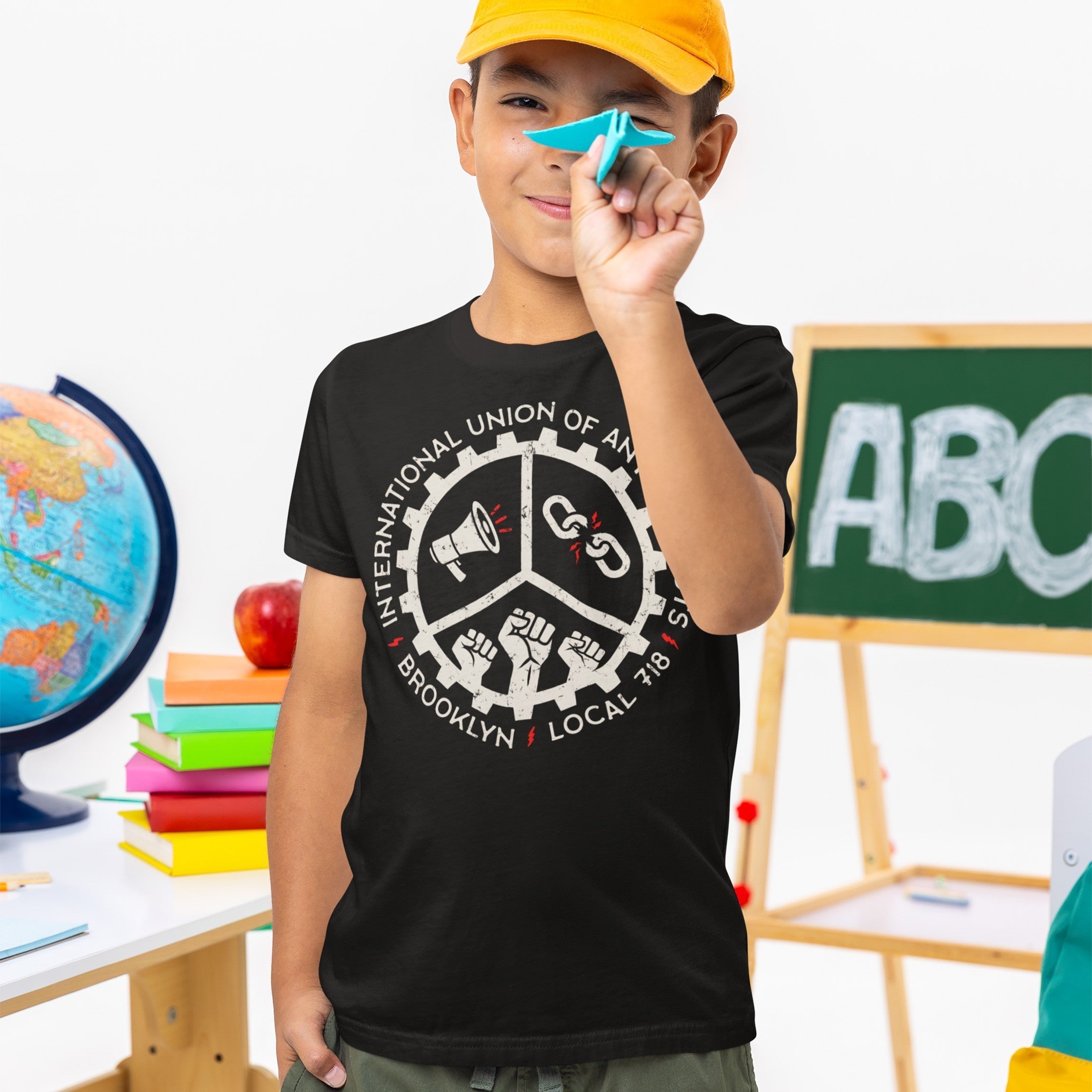 Young boy holds a blue paper airplane while wearing the Brooklyn Anti-Fascist Union T-shirt featuring a circular logo with a megaphone, broken chain, and raised fists, symbolizing solidarity and anti-fascism.