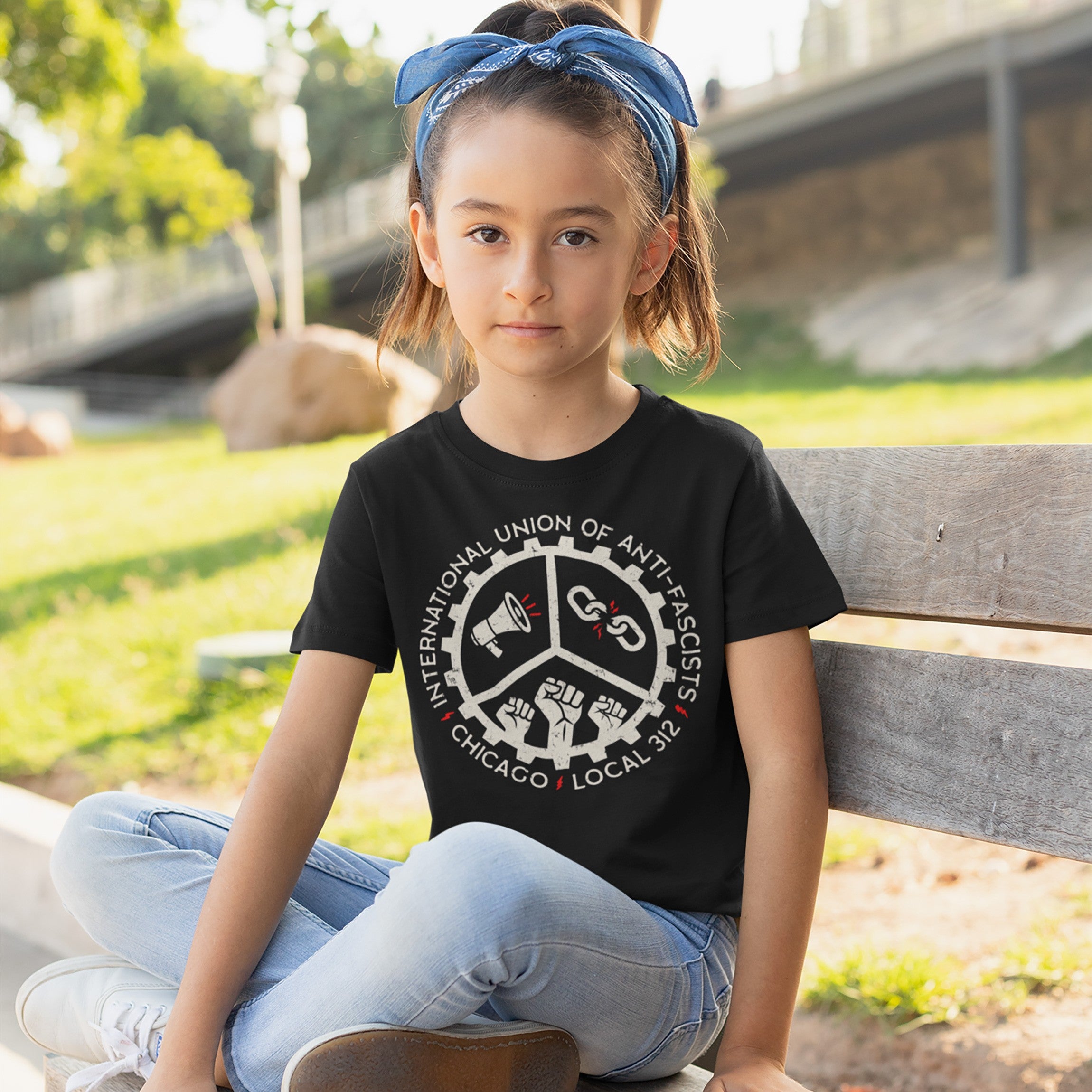 Young girl wearing the Custom Anti-Fascist Union Kids T-shirt, featuring a gear-style logo with a megaphone, broken chain, and fists, sitting on a park bench, reflecting activism and empowerment.