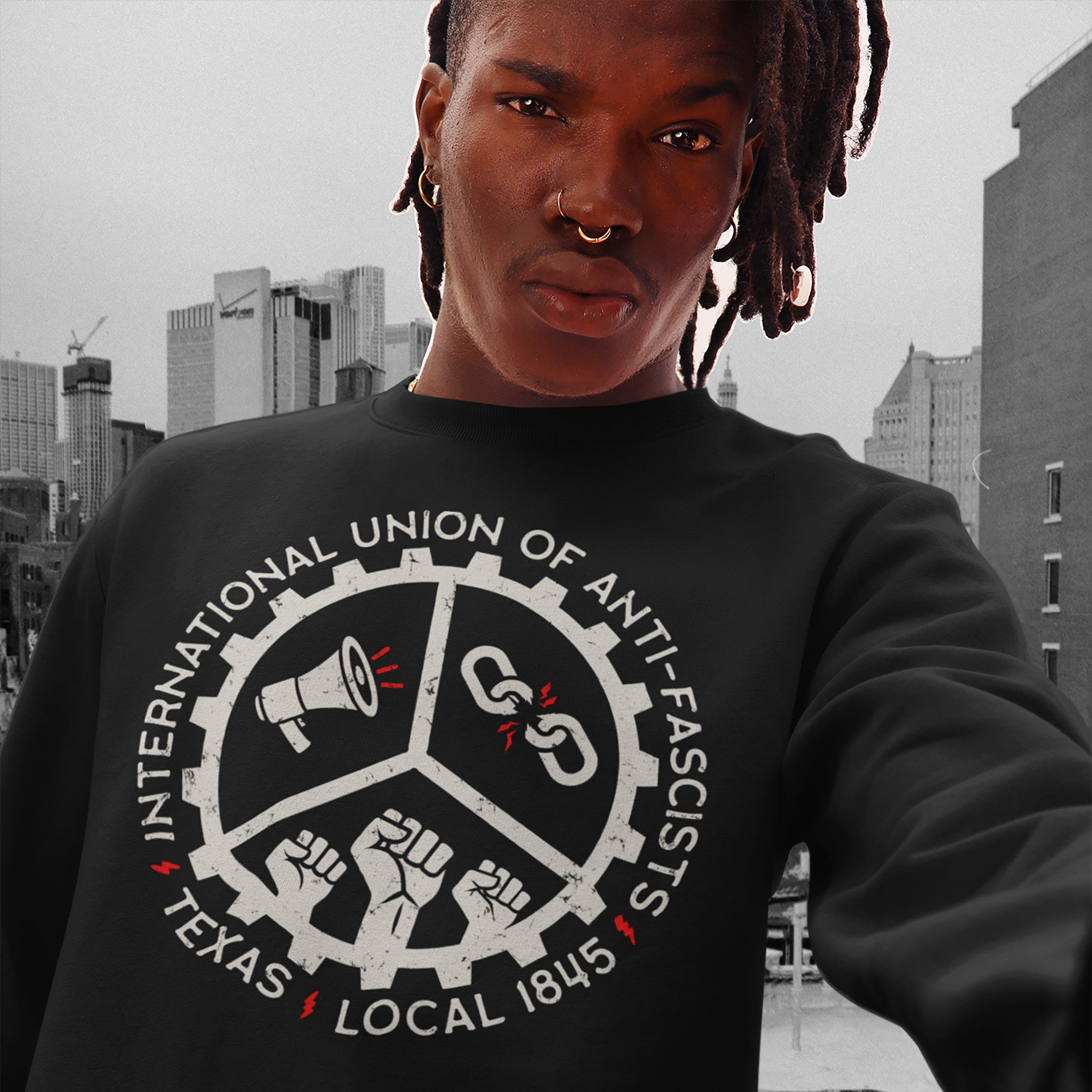 Person wearing Texas Anti-Fascist Union Hoodie with cog-wheel design featuring icons of a megaphone, broken chain, and raised fists, standing against a city skyline.