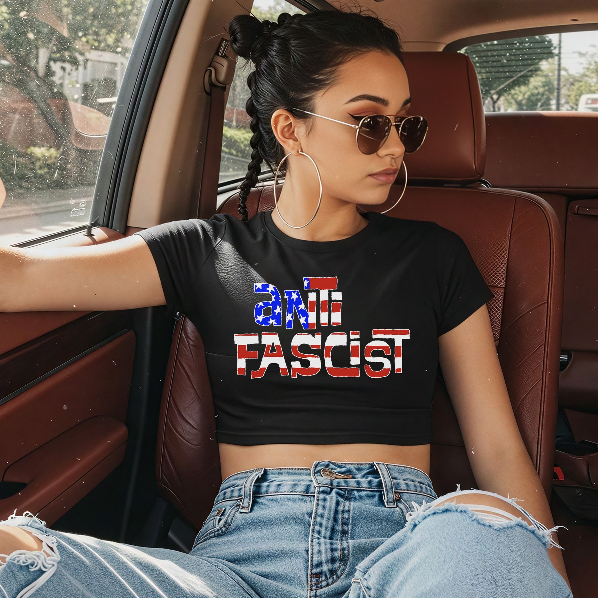 Young woman in aviator sunglasses and hoop earrings wearing Anti-Fascist USA Women's Baby Tee, a black cropped shirt with American-flag motif and “ANTIFASCIST” text, styled with light-wash ripped jeans.
