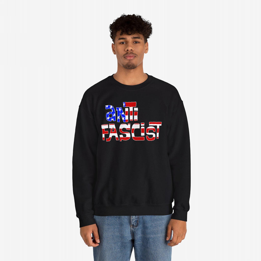 Young man wearing the Anti Fascist USA Shirt, a black crewneck sweatshirt with anti fascist in American-flag patterned letters, paired with light-wash blue jeans, symbolizing resistance and patriotism.