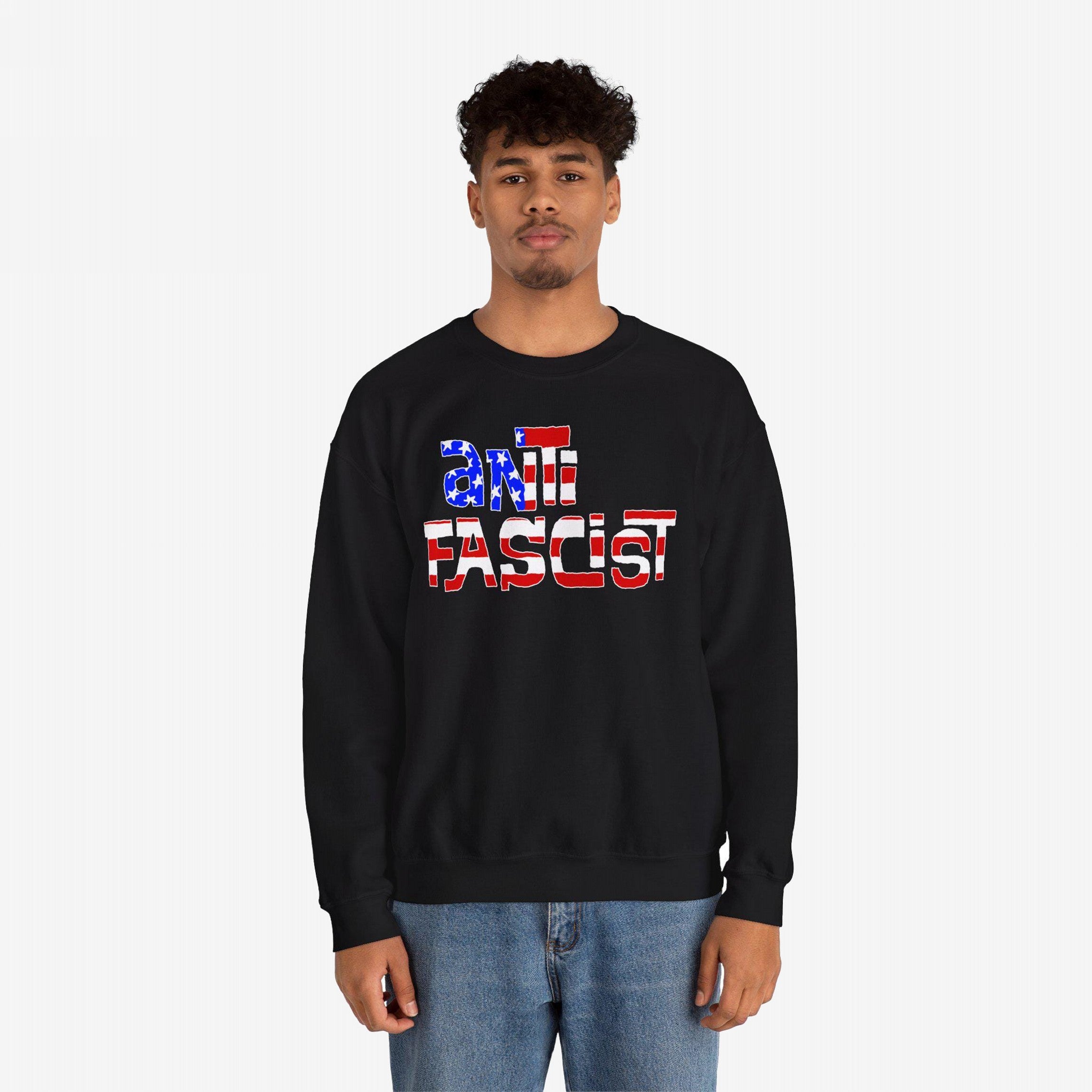 Young man wearing the Anti Fascist USA Shirt, a black crewneck sweatshirt with anti fascist in American-flag patterned letters, paired with light-wash blue jeans, symbolizing resistance and patriotism.