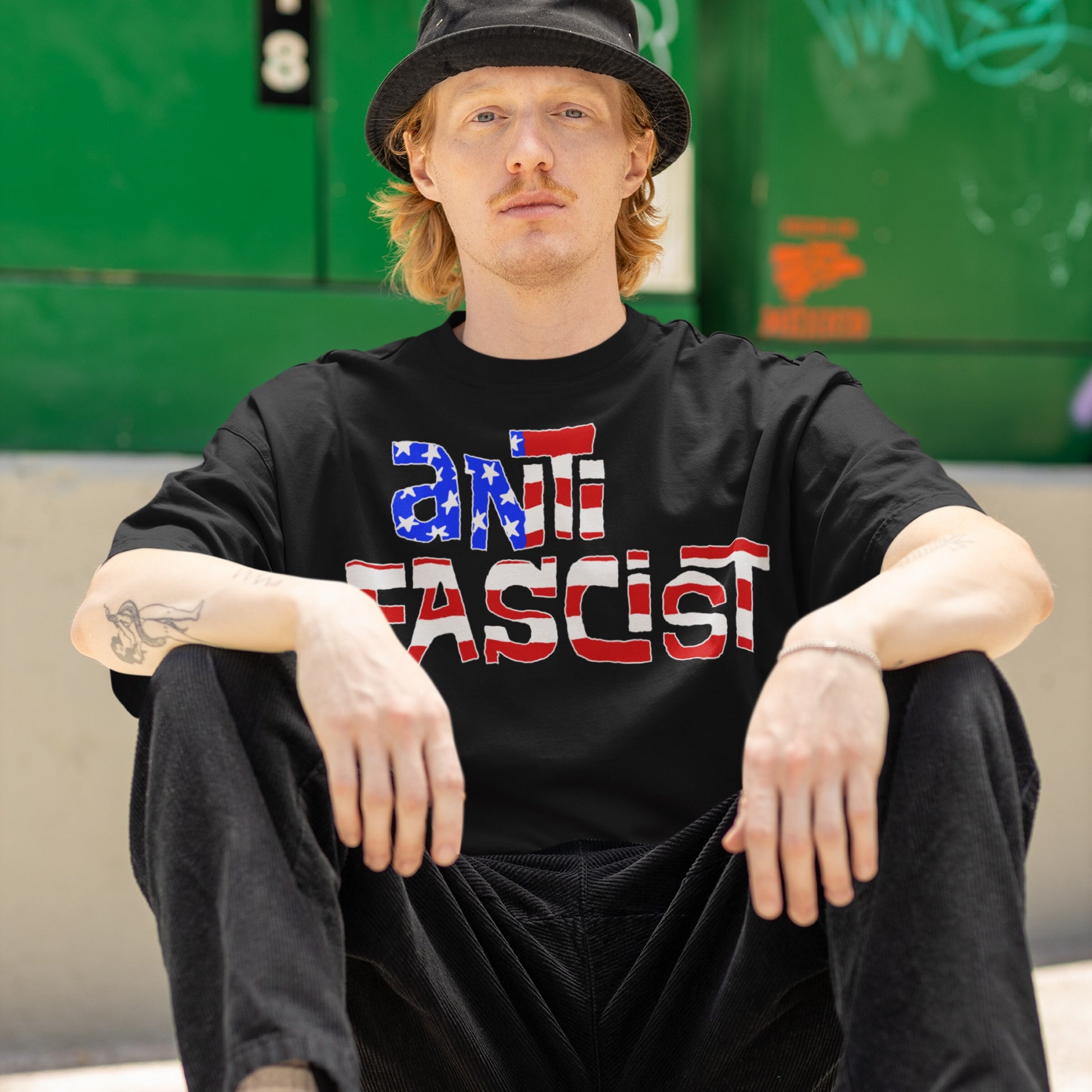 Young man in black bucket hat wears Anti-Fascist USA T-shirt, featuring a bold red, white, and blue design, seated casually in front of a green backdrop, embodying resistance and equality.