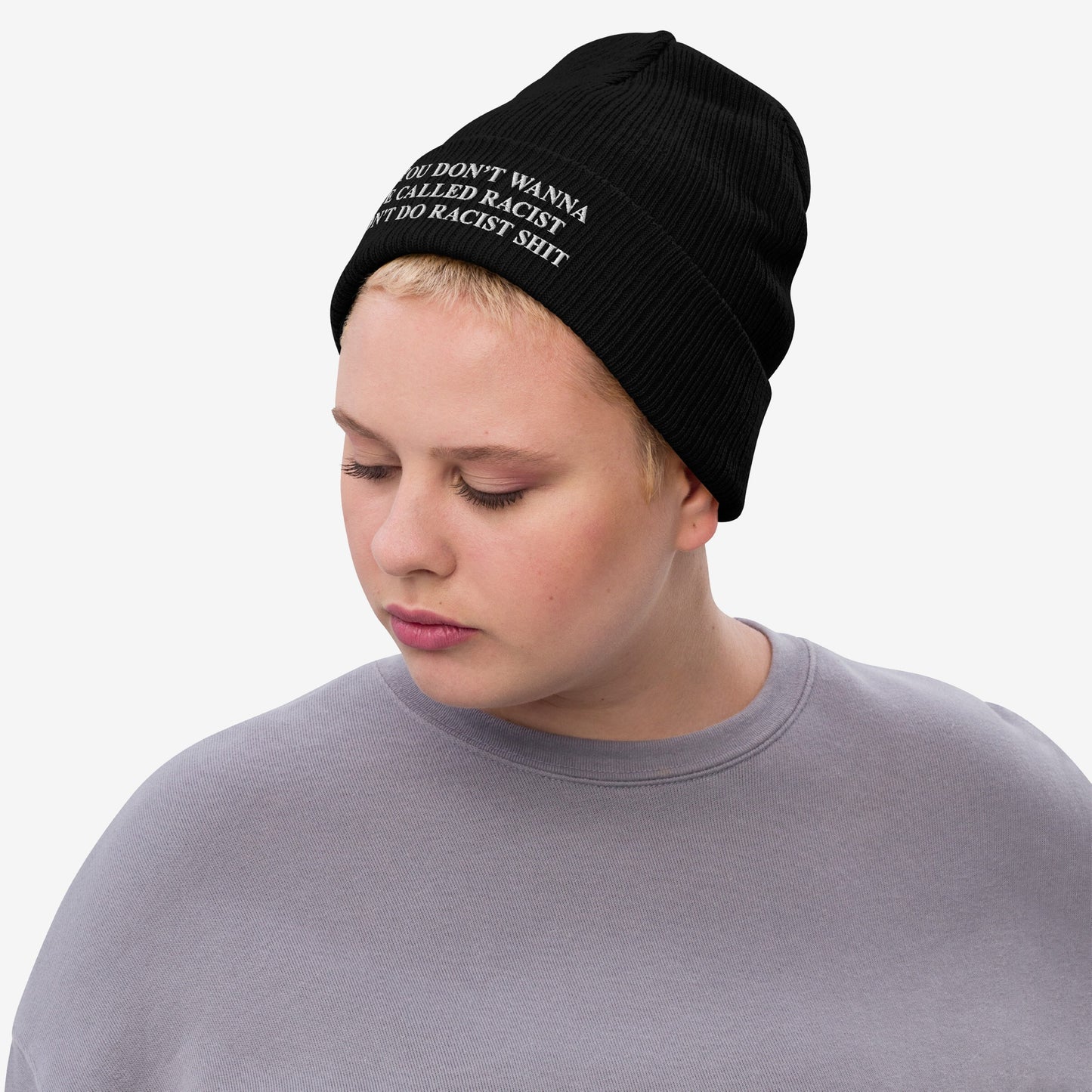 Person wearing the Anti-Racist Hat, a black knit beanie with a bold message confronting racism, paired with a light-gray crewneck. The wearer is looking down, emphasizing the hat's impactful statement.