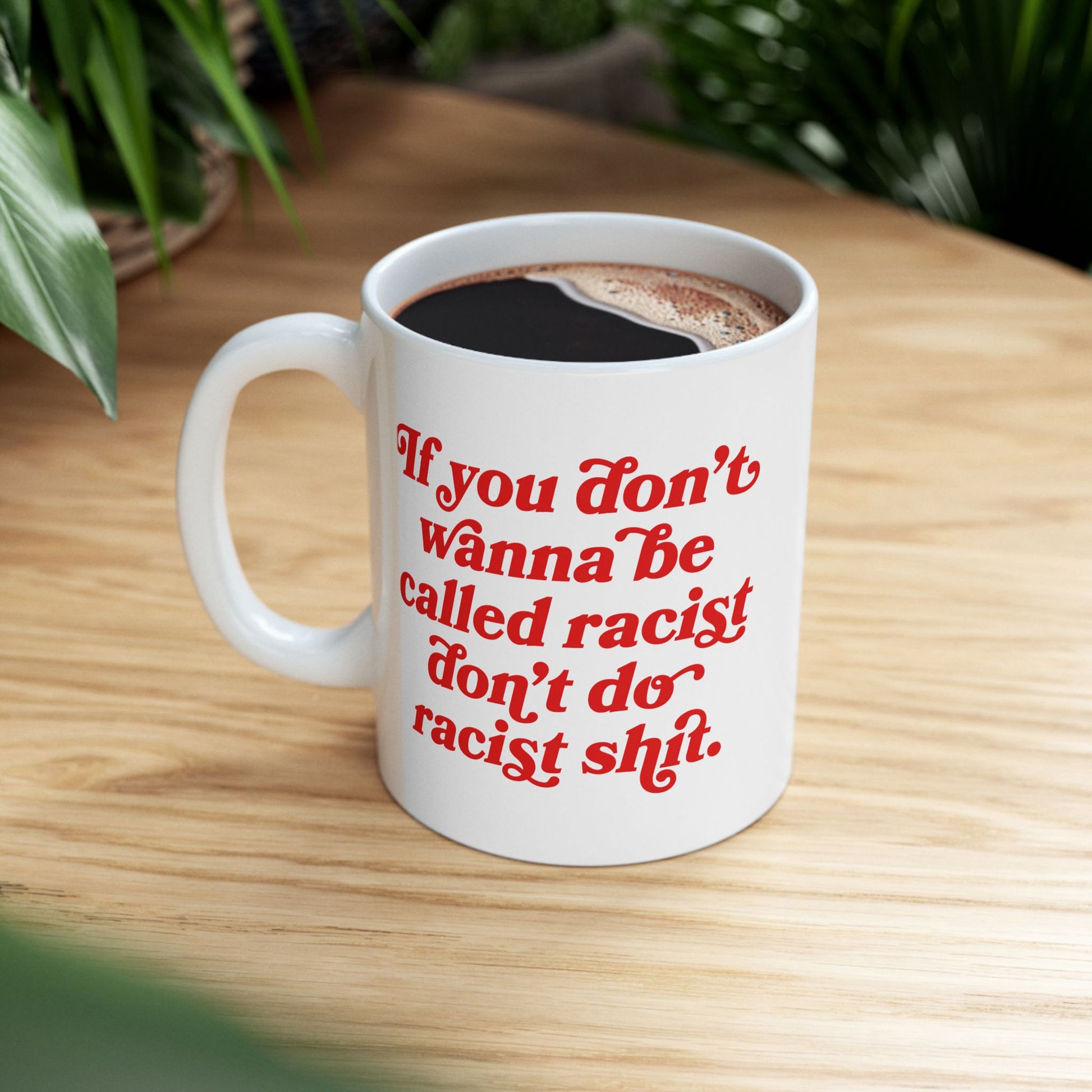Anti-Racist Mug on a wood tabletop, filled with black coffee, features bold red script: If you don’t wanna be called racist don’t do racist shit, embodying a strong message against complacency.