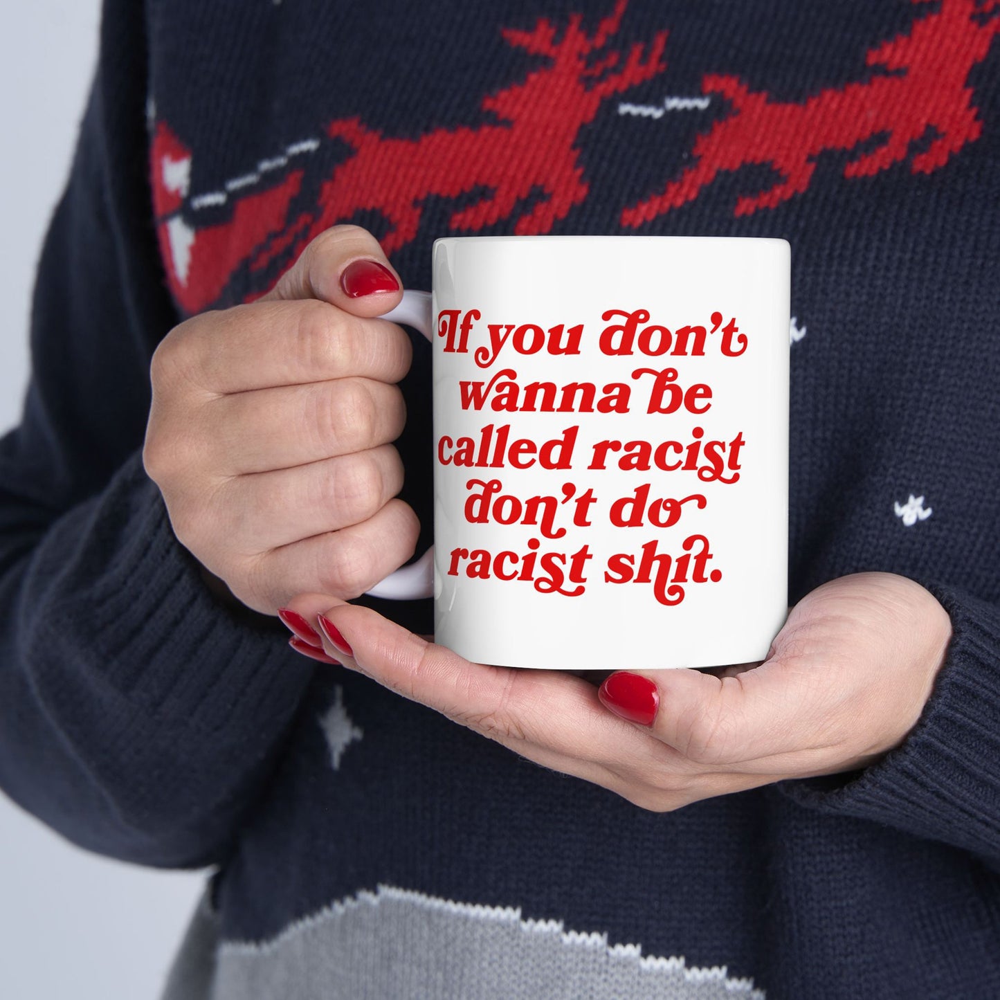 Close-up of the Anti-Racist Mug featuring bold red text, If you don’t wanna be called racist don’t do racist shit, held by a person in a patterned sweater with red nails.