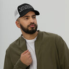 A young man models the Anti-Racist Hat, featuring a bold slogan on a black front panel, emphasizing action against racism. He wears a white T-shirt and olive jacket, embodying a strong stance.