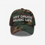 Art Drugs Music Life Hat: A woodland camo baseball cap featuring bold white embroidery of ART DRUGS MUSIC LIFE, symbolizing a creative, unapologetic spirit. Sturdy cotton with a curved brim and adjustable fit.