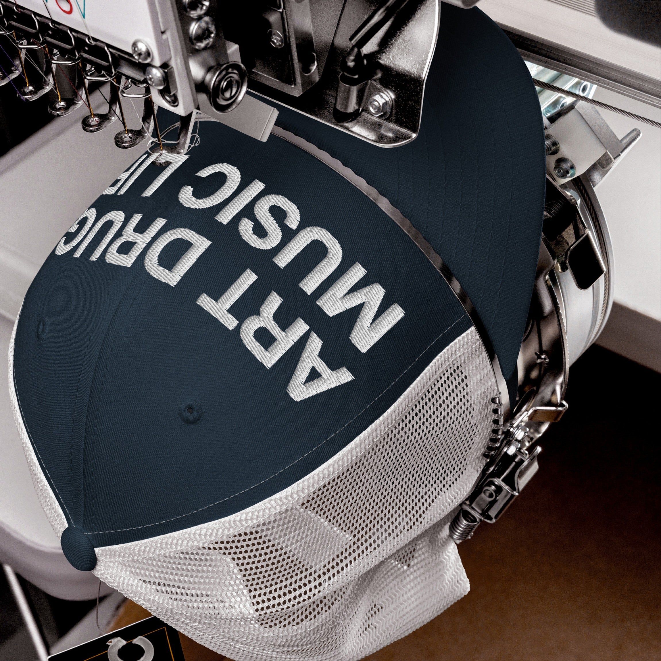 Art Drugs Music Life Hat in an embroidery machine. The navy trucker cap showcases bold white lettering, embodying a creative manifesto for those who embrace art, chaos, and sound.