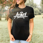 Person outdoors wearing the Atlanta Handstyle Women's Baby Tee, featuring a graffiti-style ATLANTA logo with a halo. Paired with light-wash jeans, this look embodies the city's vibrant, rebellious spirit.