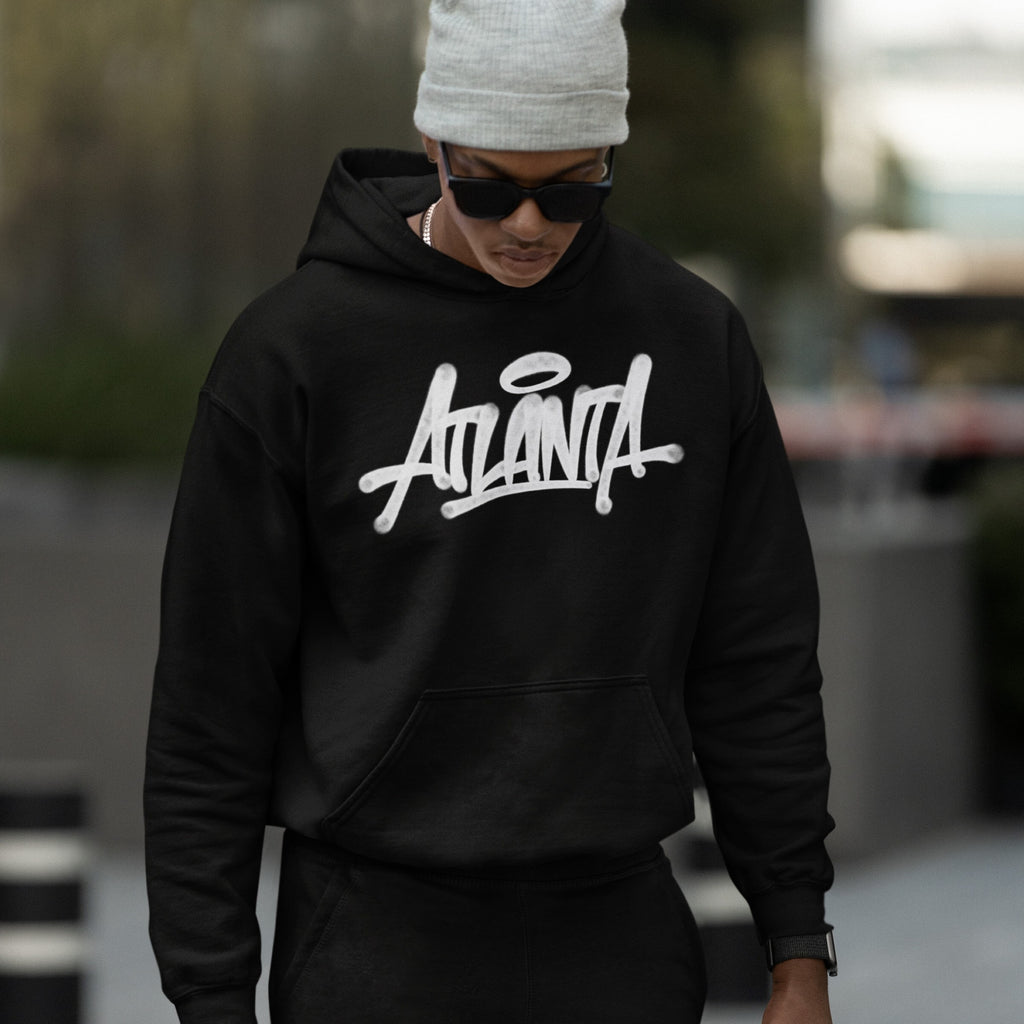 Young person in an Atlanta Handstyle Hoodie, showcasing bold ATL pride with stylized script, complemented by a gray beanie and sunglasses, embodying urban streetwear vibes in an out-of-focus urban setting.