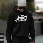 Young person in an Atlanta Handstyle Hoodie, showcasing bold ATL pride with stylized script, complemented by a gray beanie and sunglasses, embodying urban streetwear vibes in an out-of-focus urban setting.