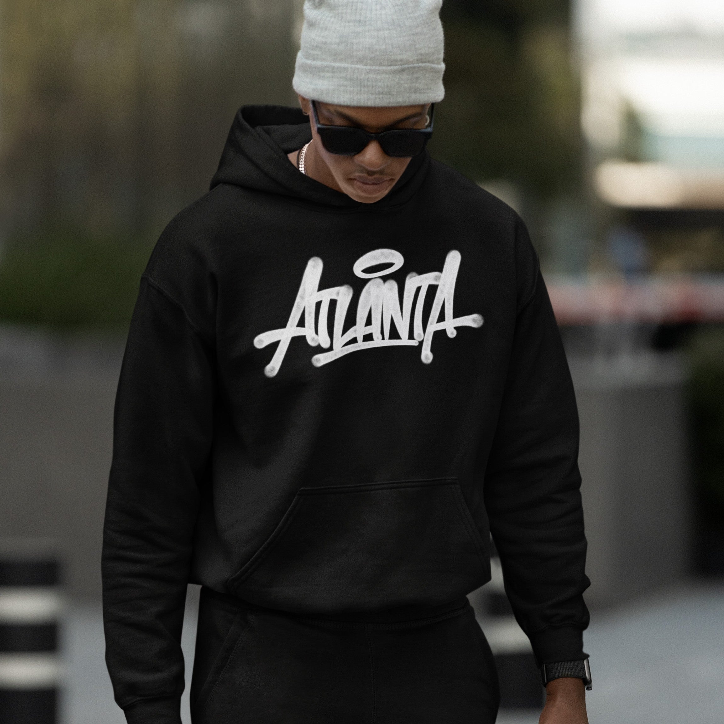 Young person in an Atlanta Handstyle Hoodie, showcasing bold ATL pride with stylized script, complemented by a gray beanie and sunglasses, embodying urban streetwear vibes in an out-of-focus urban setting.