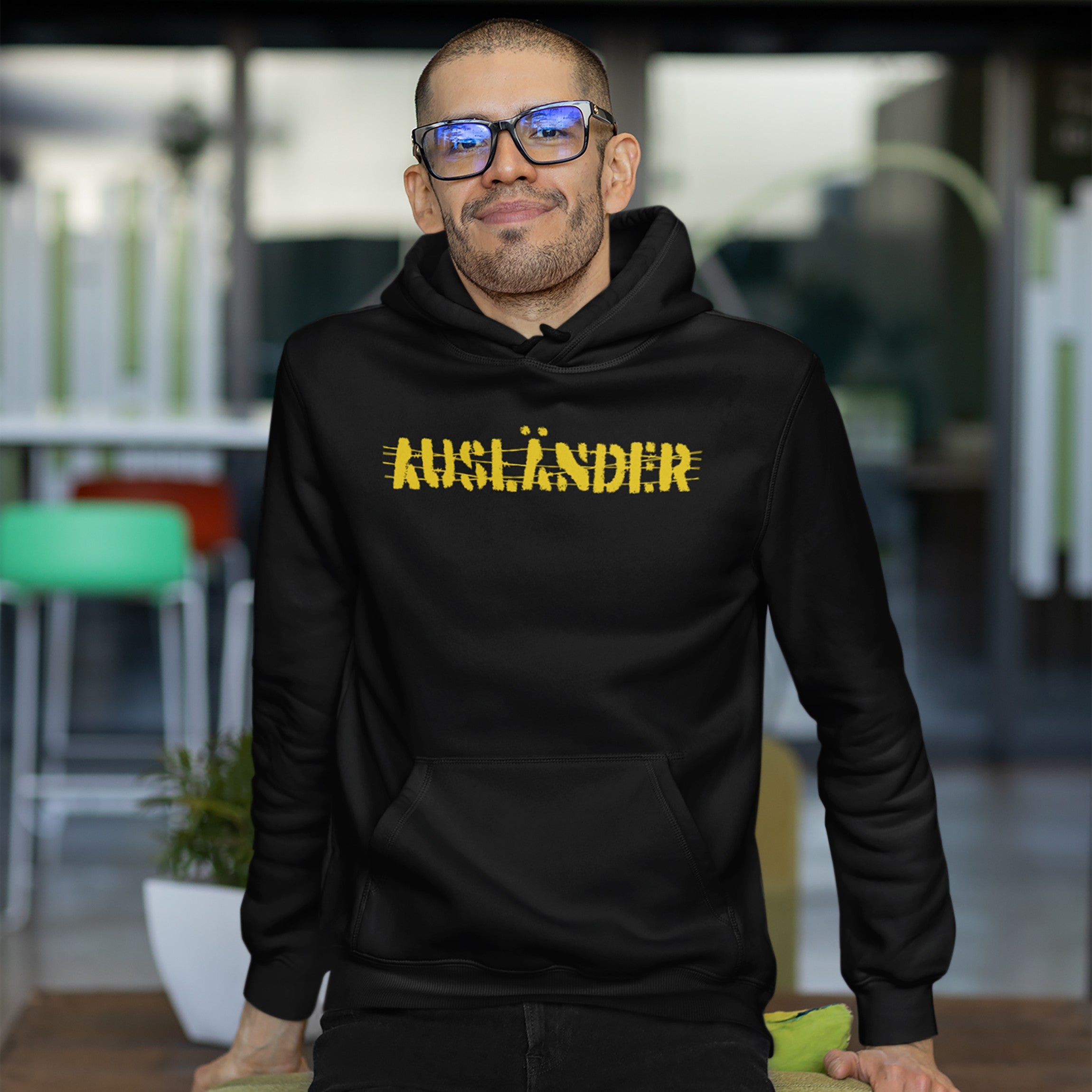 Man wearing Ausländer Hoodie with AUSLÄNDER in distressed yellow, embodying resilience and empowerment in a modern office space. Perfect for those reclaiming identity and showcasing strength.