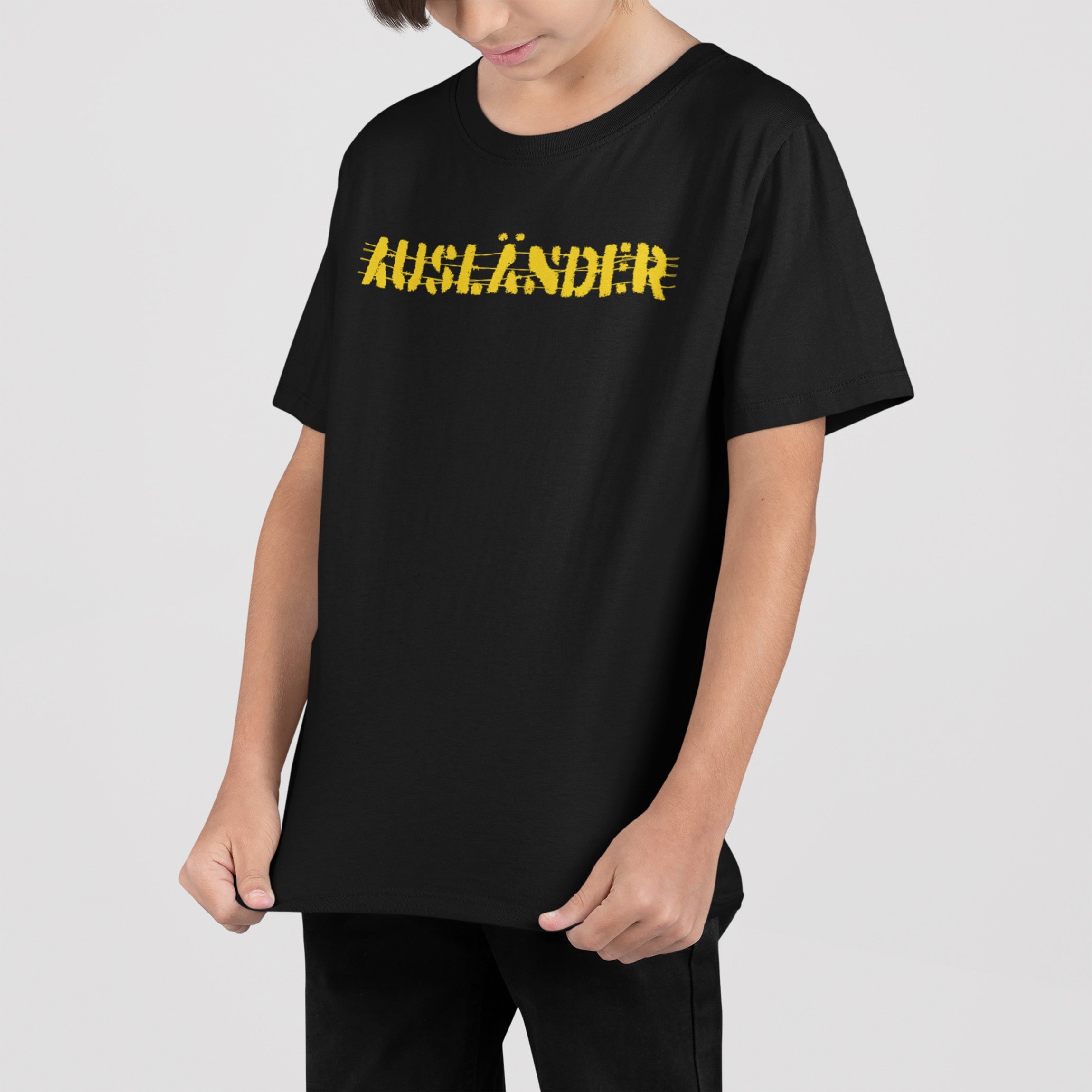Child wearing Ausländer T-shirt with bold yellow text and strike-throughs, holding the hem and looking down, symbolizing resistance and empowerment against borders. Available in various styles and sizes.