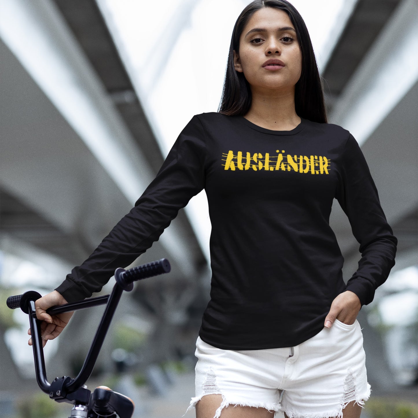 Young woman in urban walkway wearing the Ausländer T-shirt, a black long-sleeved top with a bold yellow print, symbolizing resistance and solidarity, paired with white distressed denim shorts, holding a bicycle.