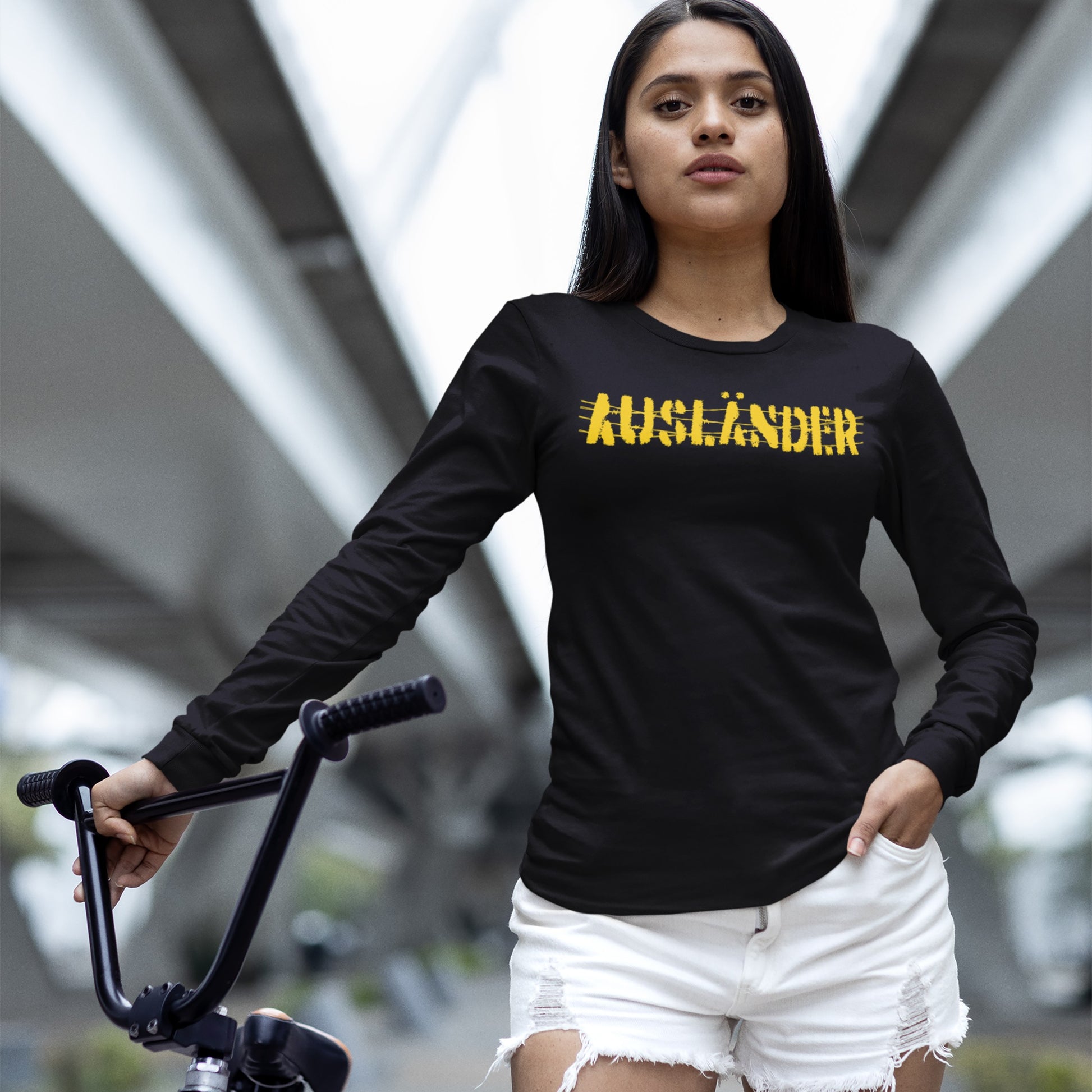 Young woman in urban walkway wearing the Ausländer T-shirt, a black long-sleeved top with a bold yellow print, symbolizing resistance and solidarity, paired with white distressed denim shorts, holding a bicycle.