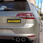 Rear view of a light-colored hatchback with an AUSLÄNDER bumper sticker in distressed yellow on black. The Auslander Sticker & Magnet symbolizes identity reclamation and solidarity against nationalism.