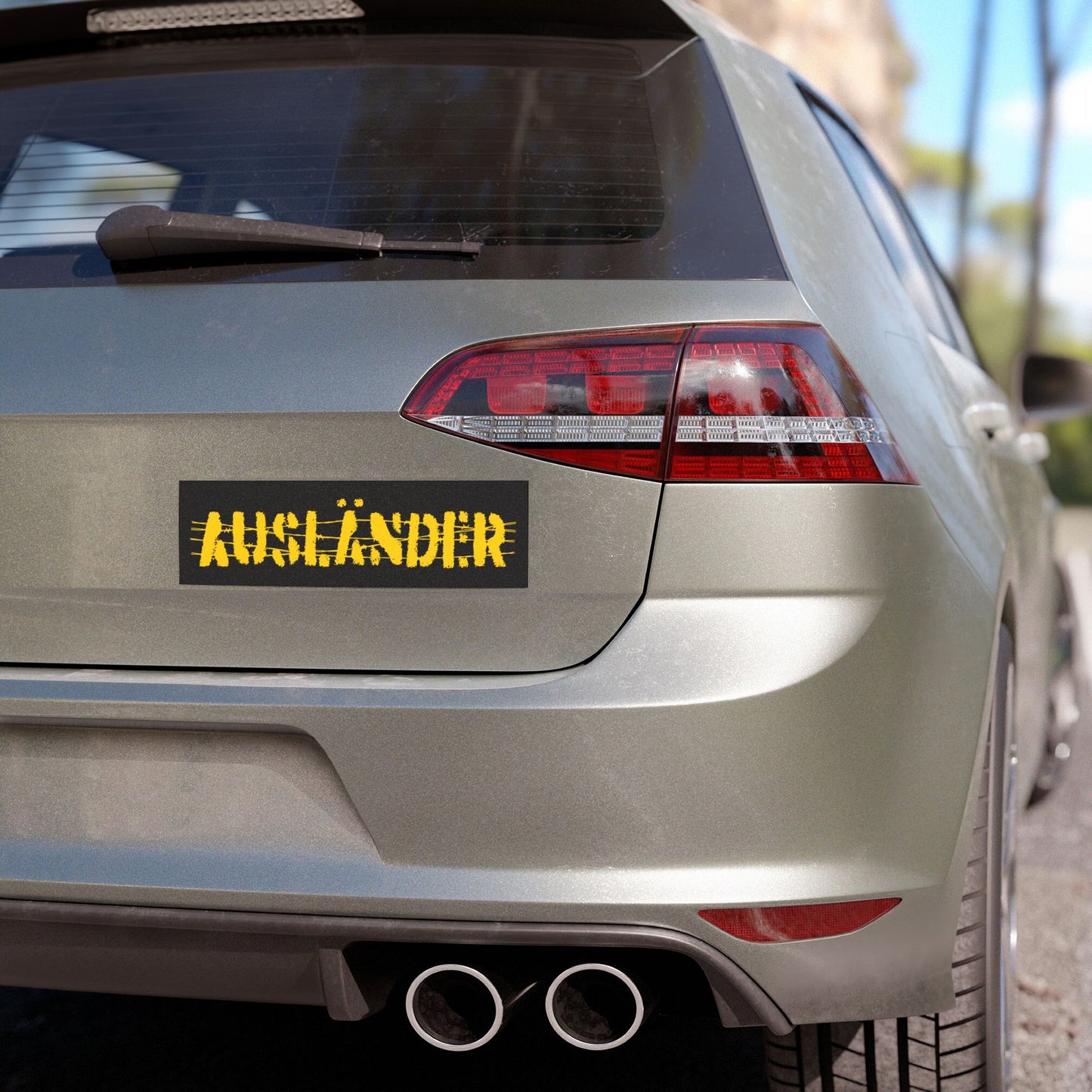 Rear view of a light-colored hatchback with an AUSLÄNDER bumper sticker in distressed yellow on black. The Auslander Sticker & Magnet symbolizes identity reclamation and solidarity against nationalism.
