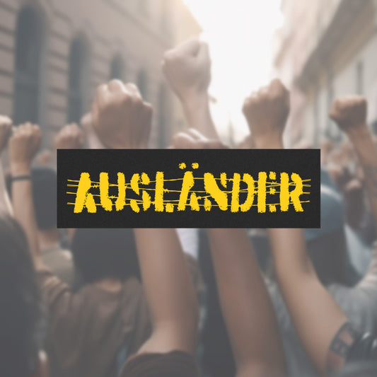 Ausländer Sticker & Magnet featuring a distressed, stencil-style AUFZÄNDER design on a black banner. The background shows a blurred crowd, symbolizing solidarity and strength against nationalism and oppression.