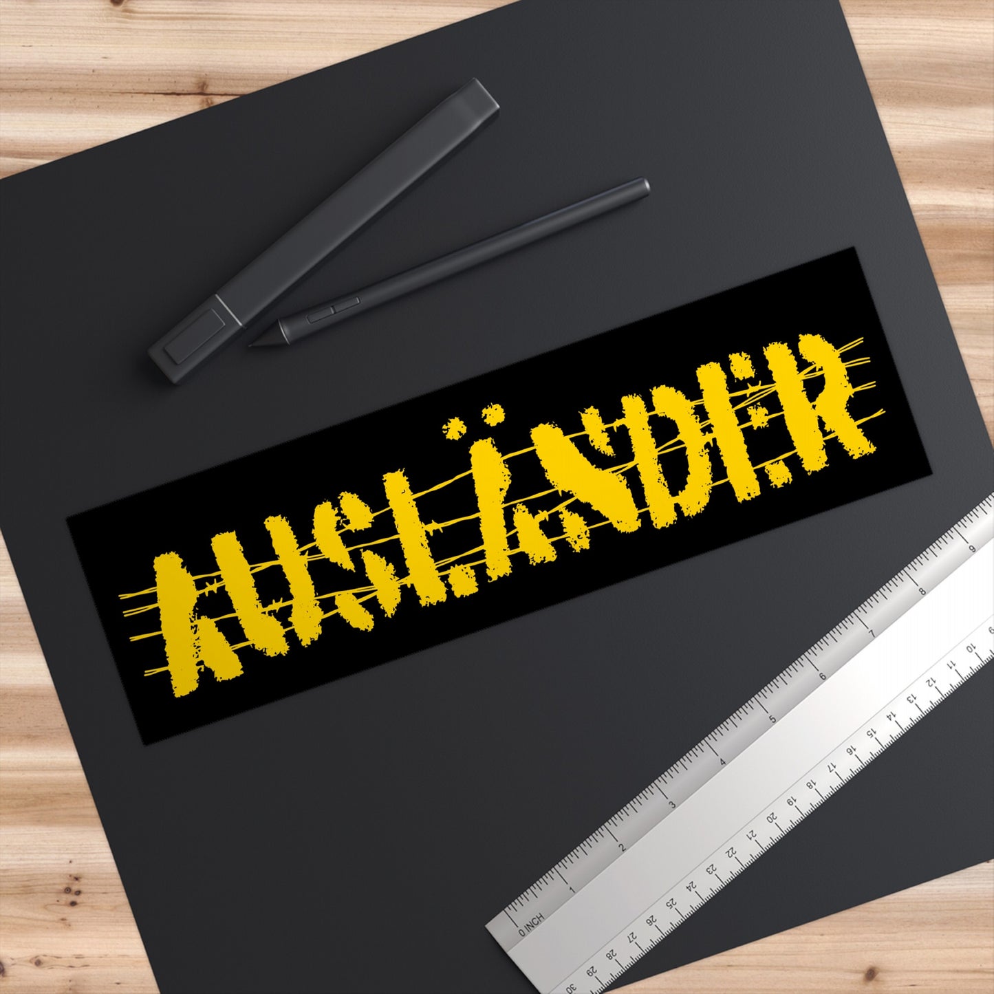 Auslander Sticker & Magnet displayed on a design desk, featuring distressed yellow barbed-wire lettering symbolizing resilience against nationalism. Set on a dark mat with a stylus, ruler, and wood tabletop.