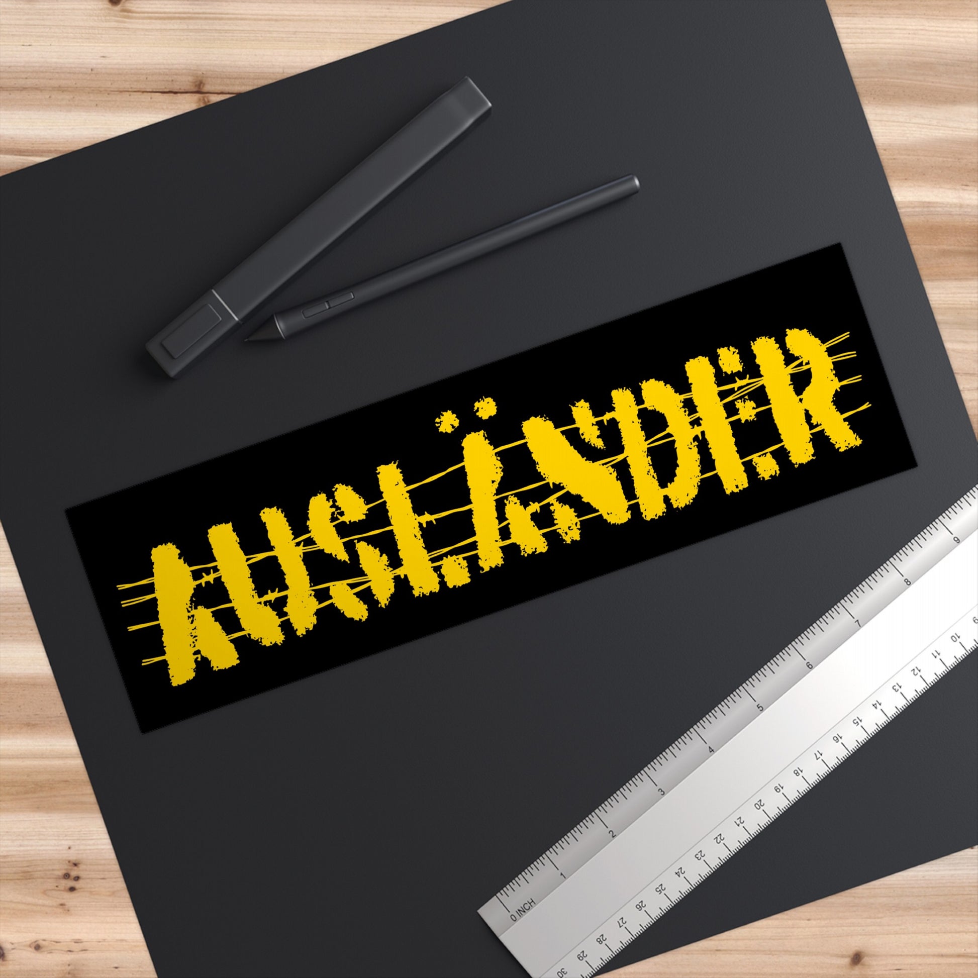Auslander Sticker & Magnet displayed on a design desk, featuring distressed yellow barbed-wire lettering symbolizing resilience against nationalism. Set on a dark mat with a stylus, ruler, and wood tabletop.