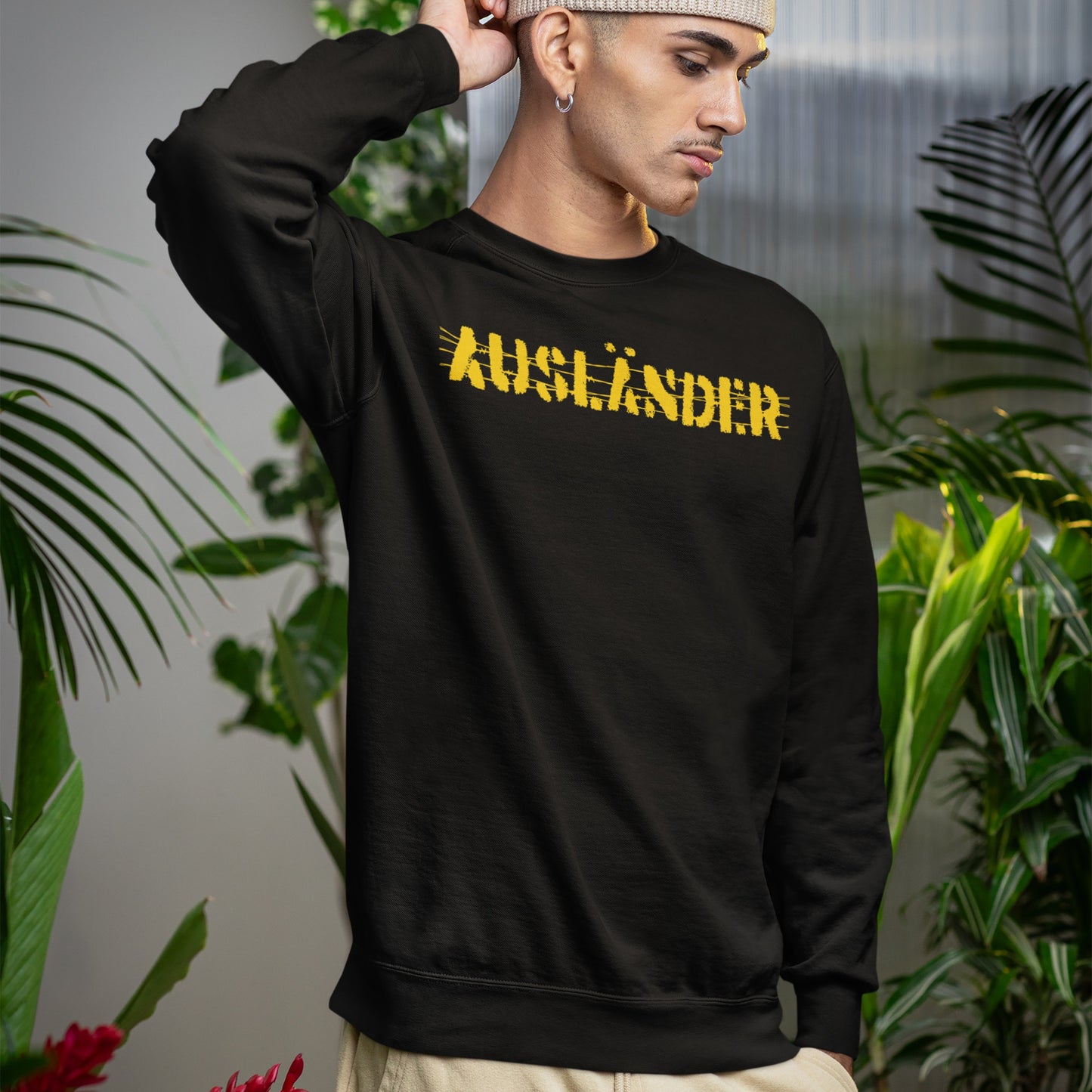 Young man in a black Ausländer crew-neck sweatshirt, featuring a bold, distressed yellow print. Surrounded by lush plants, he embodies defiant style and empowerment, reflecting a strong anti-discrimination message.