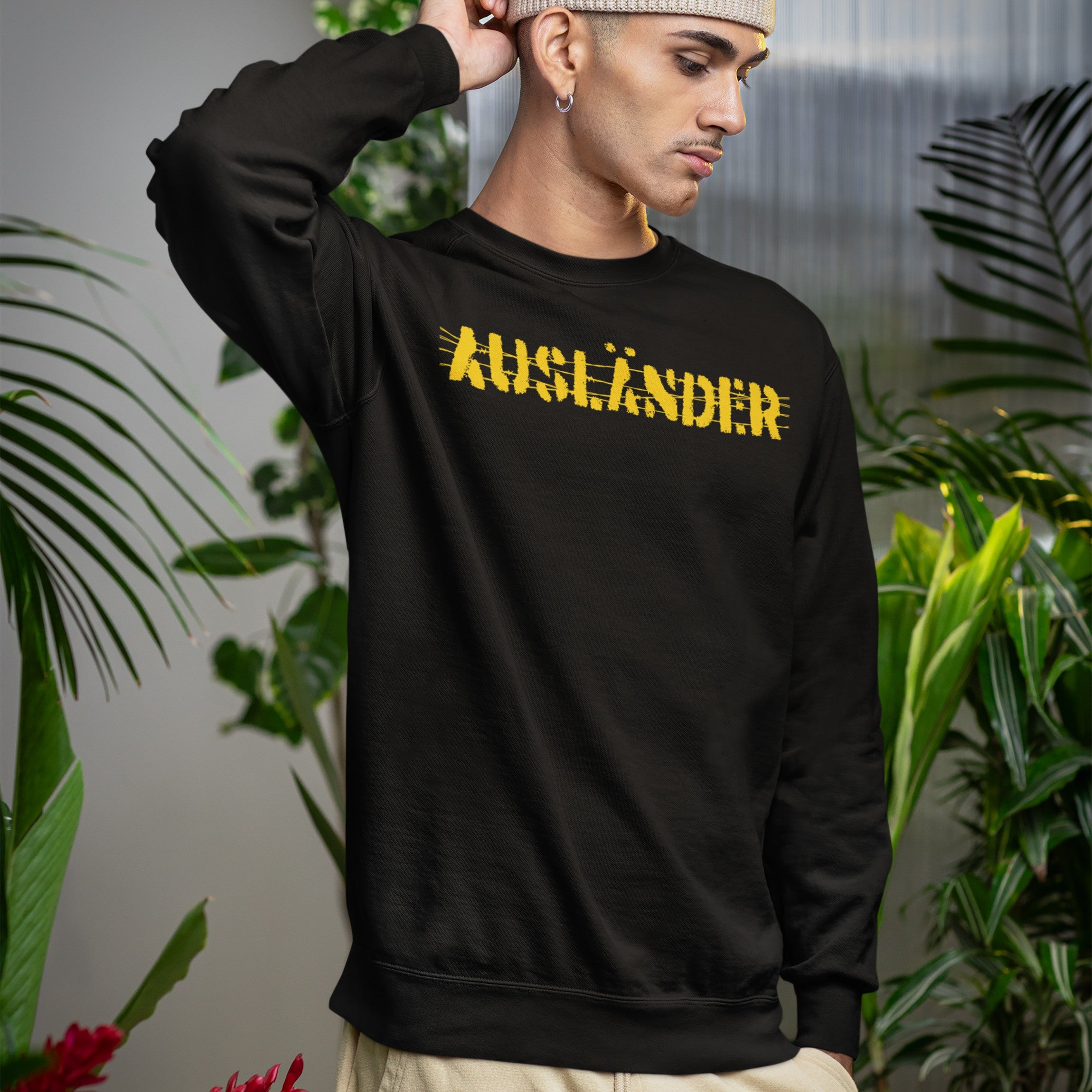 Young man in a black Ausländer crew-neck sweatshirt, featuring a bold, distressed yellow print. Surrounded by lush plants, he embodies defiant style and empowerment, reflecting a strong anti-discrimination message.