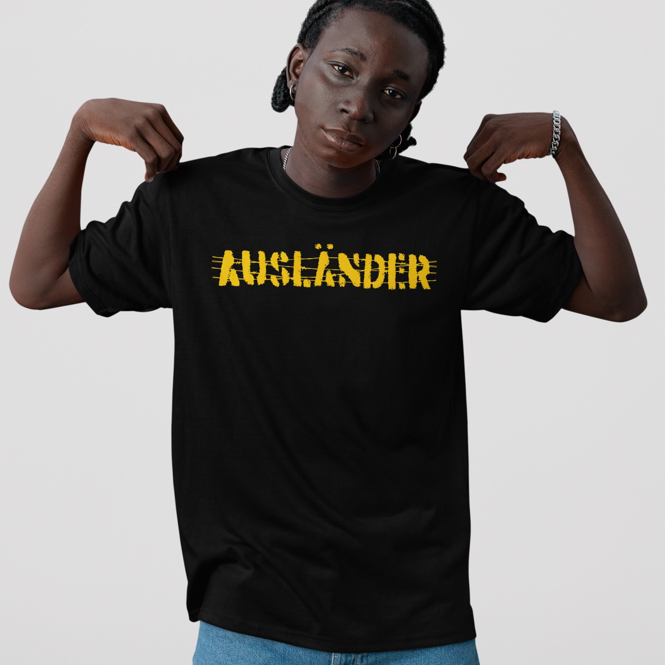Ausländer T-shirt held by person, featuring distressed yellow lettering of the word “AUSLÄNDER” scratched through, signifying defiance. Worn with braids, hoop earrings, necklace, bracelet, and blue jeans.