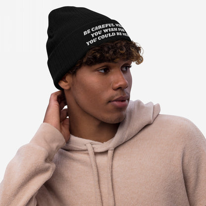 Young man in a light-beige hoodie wearing the Be Careful Hat knit beanie with bold embroidered warning text, posed against a plain background, embodying a message of resistance and style.