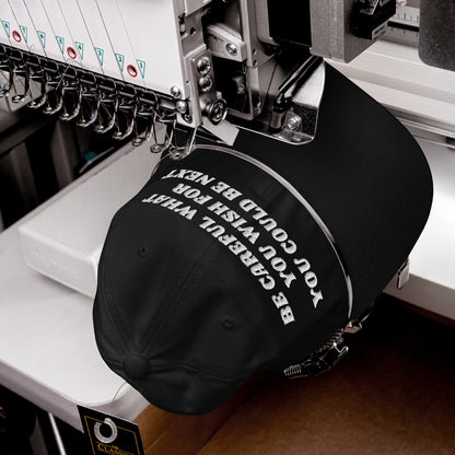 The Be Careful Hat is shown in an embroidery machine, featuring bold white stitching of its poignant message on a black cap, embodying a statement of resistance and style.