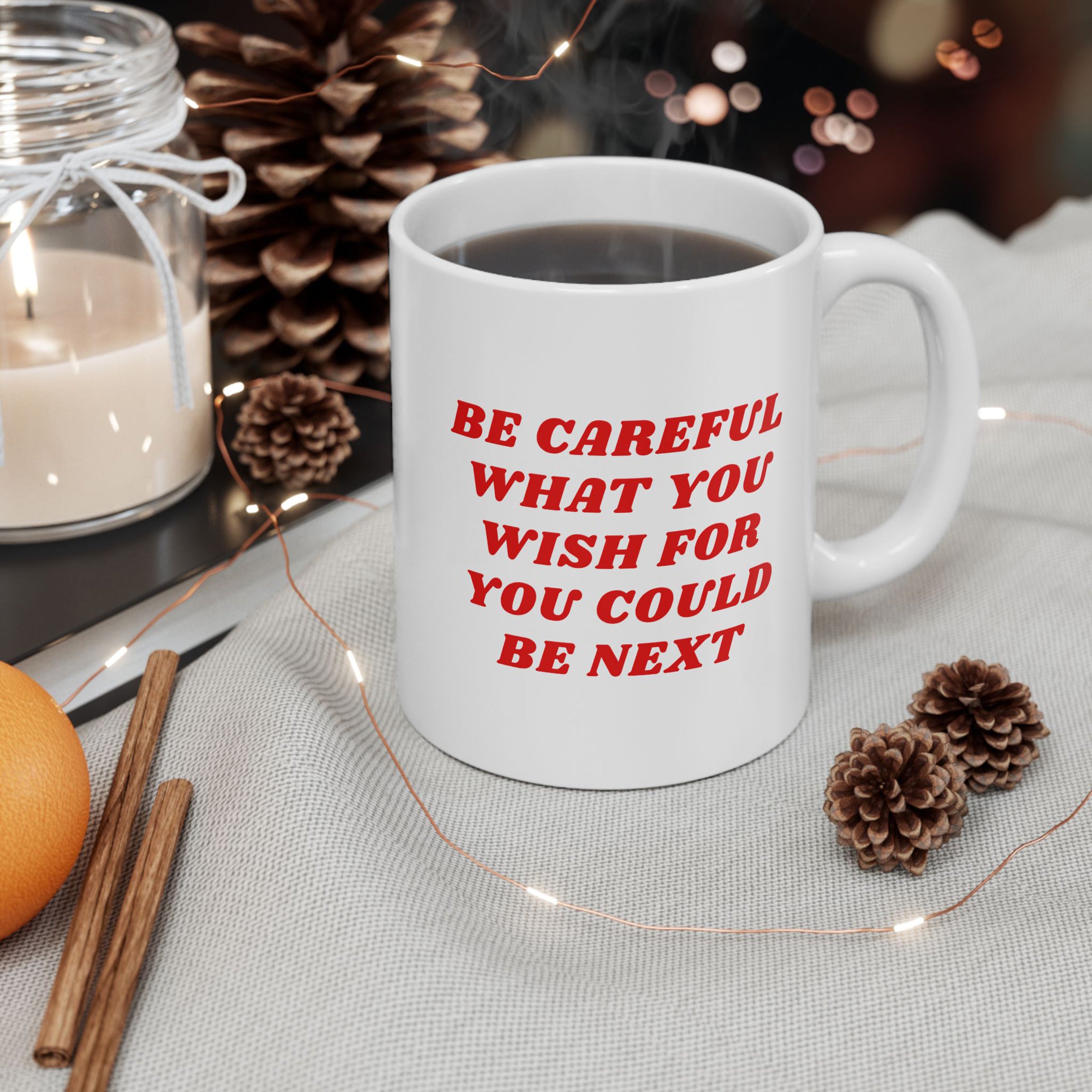 White ceramic Be Careful Mug on gray cloth, surrounded by pine cones, candle, cinnamon sticks, orange, and fairy lights. Steam rises, hinting at a hot drink inside. Cozy, rustic setting.