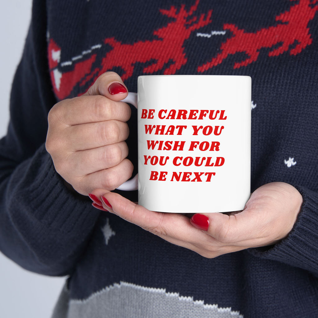 Person holding the Be Careful Mug with bold red text, red-painted nails, and a dark sweater featuring a festive pattern, reflecting a message of caution and awareness amidst societal challenges.