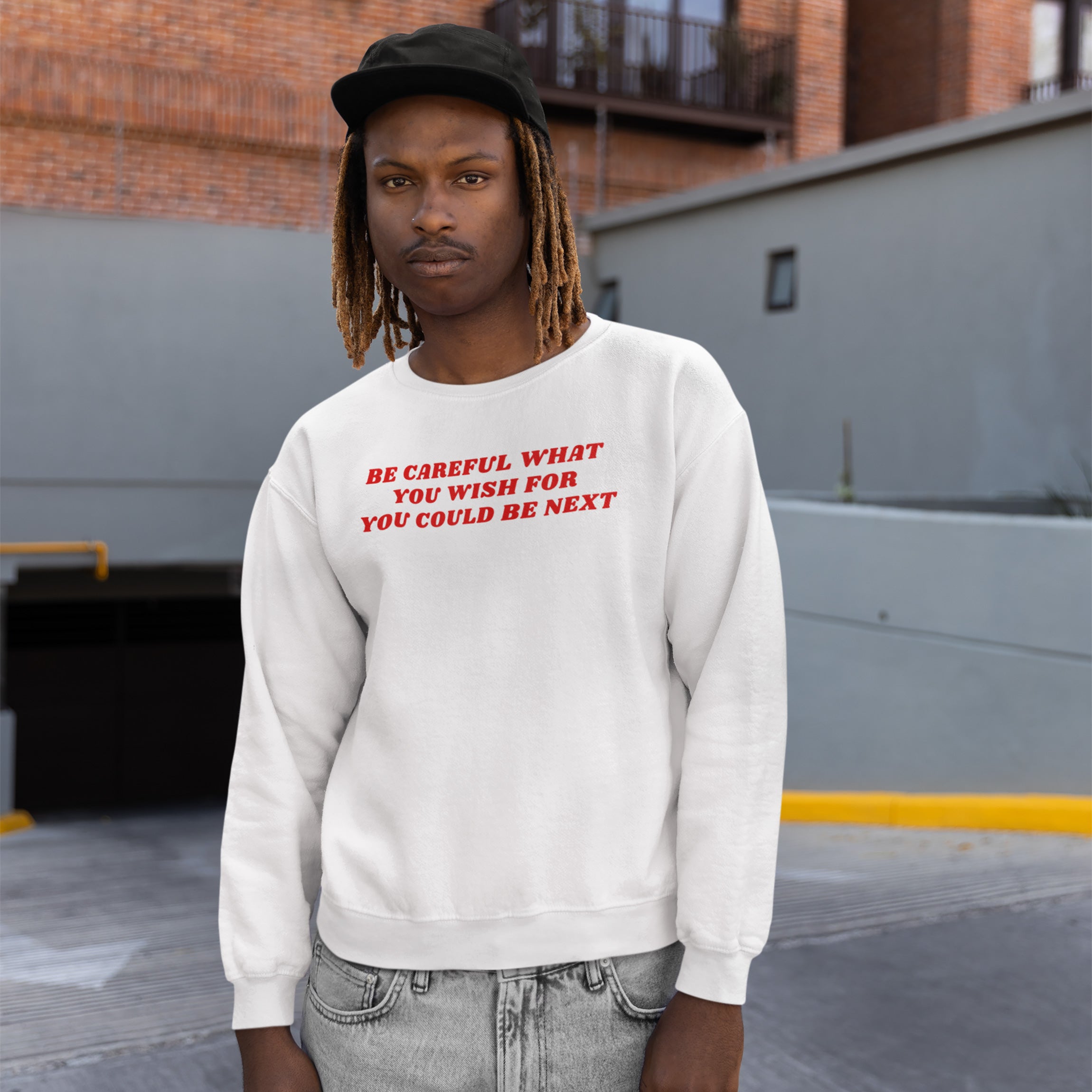 Young man in an urban setting wearing the Be Careful Shirt, a white sweatshirt with bold red text, advocating against authoritarianism and extremism, paired with light-washed gray jeans and a black baseball cap.
