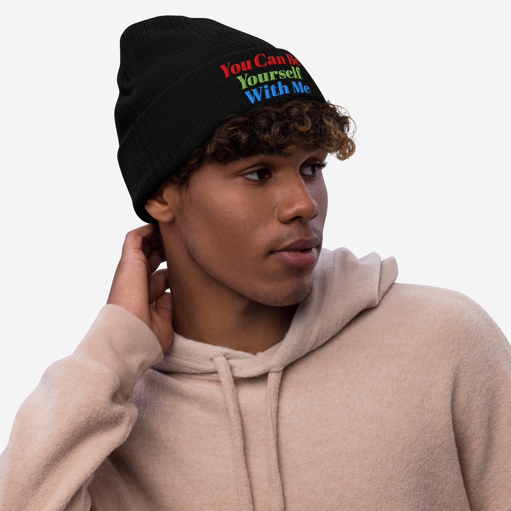 Young man in light-beige hoodie wears the Be Yourself Hat, a black knit beanie with vibrant embroidered text, You Can Be Yourself With Me, reflecting a message of inclusivity and self-expression.