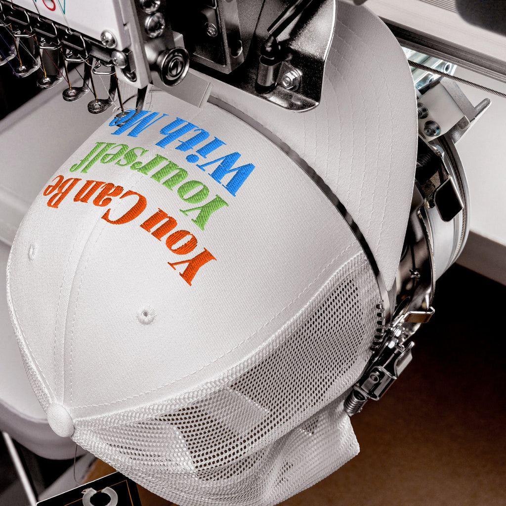 White mesh-back trucker cap under embroidery machine, stitching the phrase “You Can Be Yourself With Me” in vibrant colors on the front, embodying individuality and inclusivity. The Be Yourself Hat.