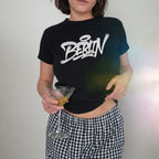 Berlin Handstyle Women's Baby Tee featuring bold graffiti-style Berlin text in white. Model wears gingham pants, holding a martini glass, embodying urban streetwear and rebellious spirit.