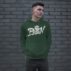 Young man in a forest-green Berlin Handstyle Hoodie stands against a gray brick wall, embodying street art culture. His hands rest in the hoodie’s pouch, gazing sideways, capturing urban boldness.