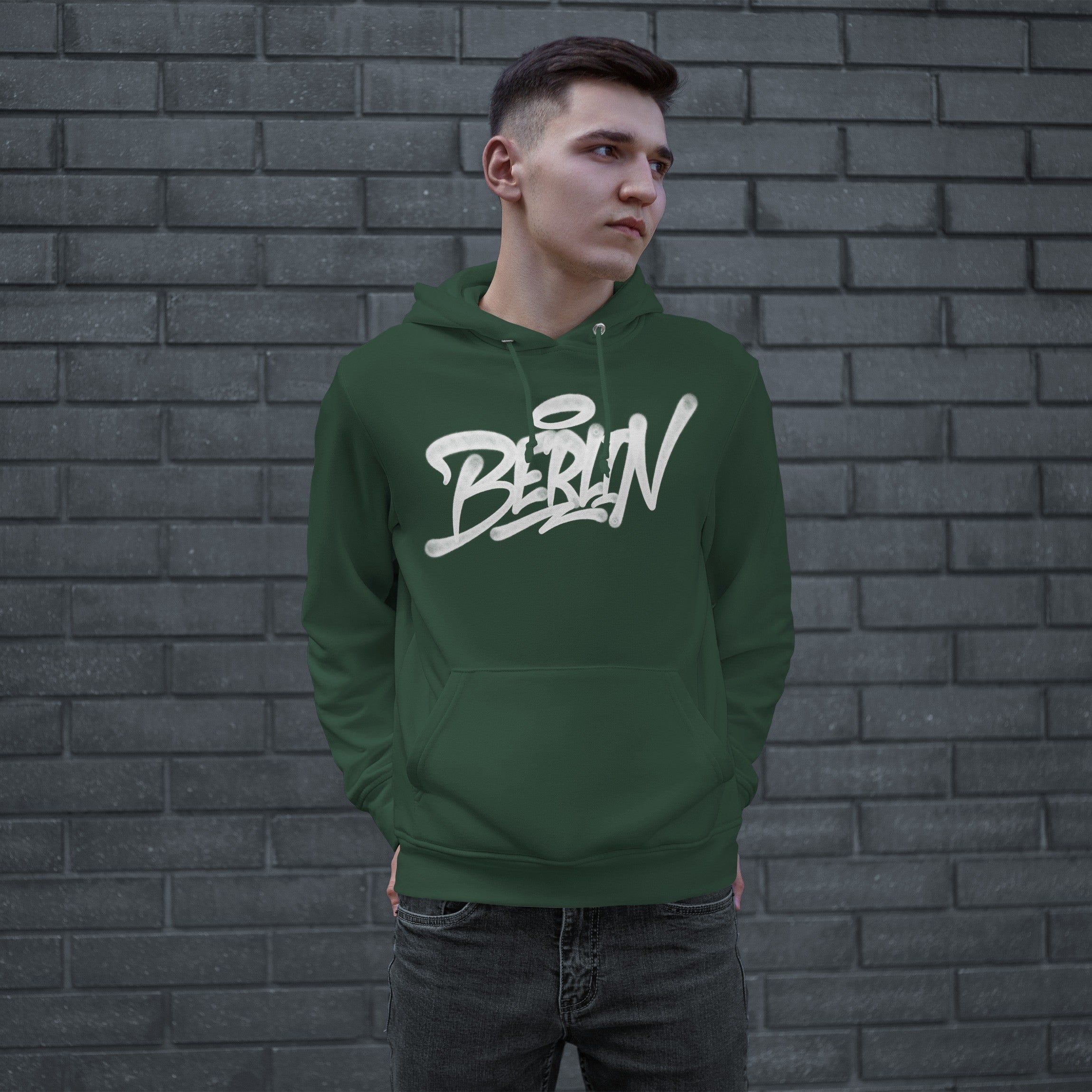 Young man in a forest-green Berlin Handstyle Hoodie stands against a gray brick wall, embodying street art culture. His hands rest in the hoodie’s pouch, gazing sideways, capturing urban boldness.