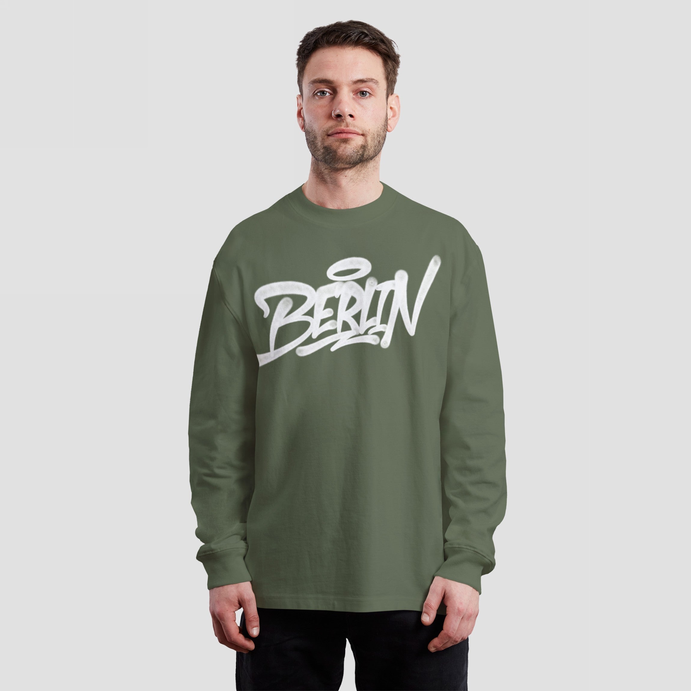 Young adult male wears the Berlin Handstyle T-shirt, featuring bold graffiti-style BERLIN text in white on an olive-green long-sleeve shirt, embodying urban attitude and street art roots.