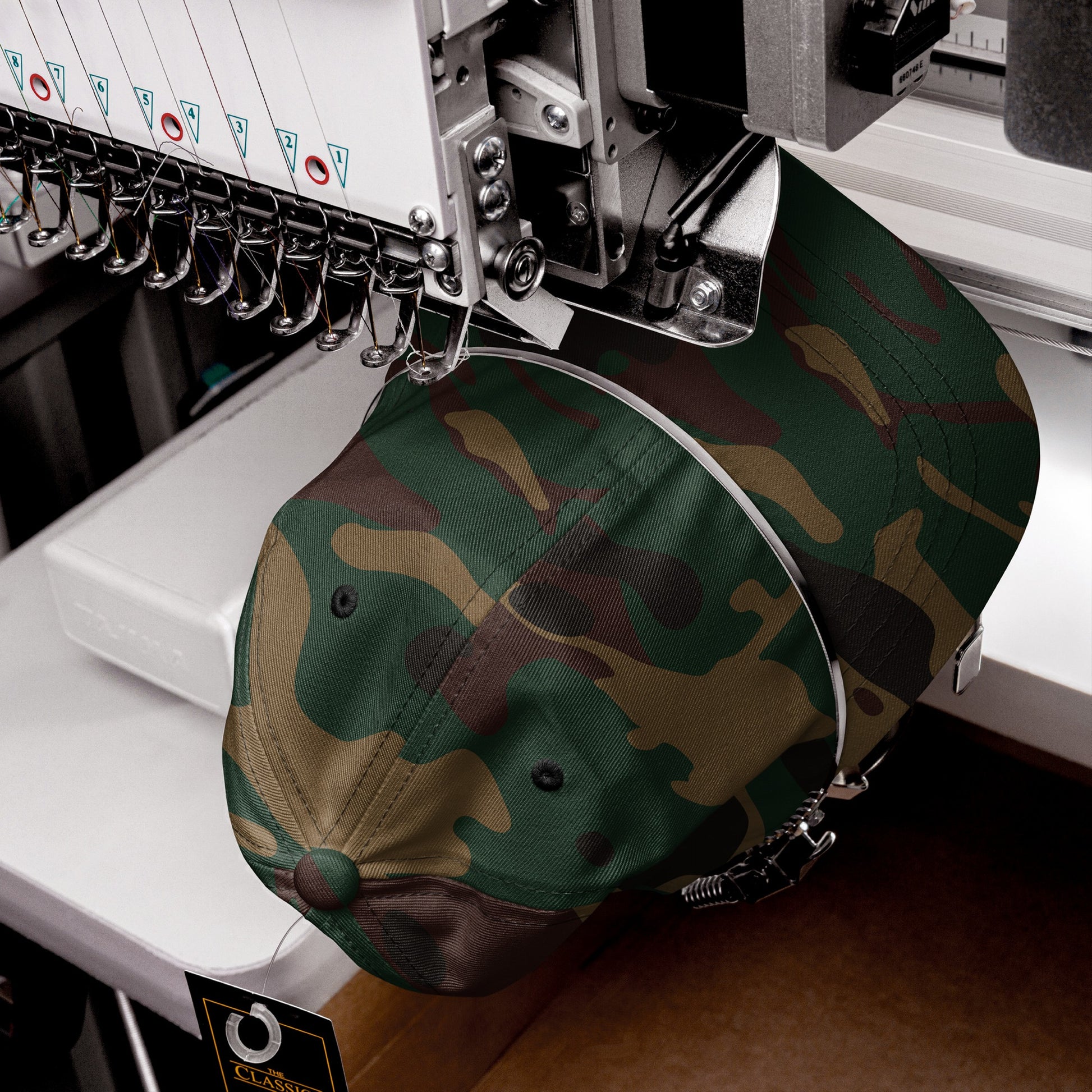 Woodland-camouflage Dad Hat In Custom Colors held in a multi-needle embroidery machine, ready for custom activist design stitching. The cap is secured by the machine’s hat frame with a Classic tag attached.