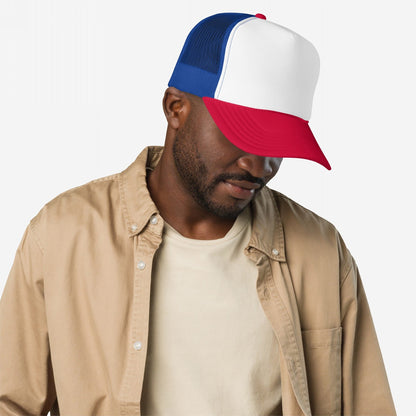 Man wearing Foam Trucker Hat In Custom Colors with white front, blue mesh, and red brim, embodying vintage activism style. He wears a beige T-shirt and tan shirt, looking relaxed.