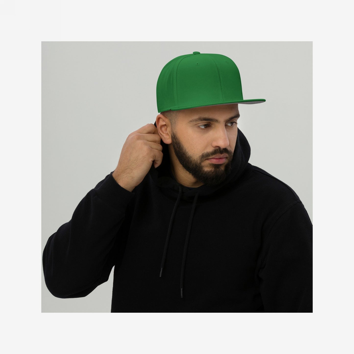 Bearded man wearing a Snapback Hat In Custom Colors, featuring a high-profile crown and flat brim, grasping his black hoodie's hood, against a light-gray background, embodying activist spirit and bold expression.